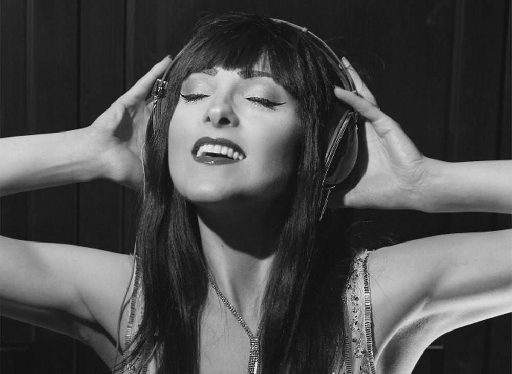 Black and white photo of a woman with closed eyes, holding headphones on her head, smiling with her lips slightly parted.