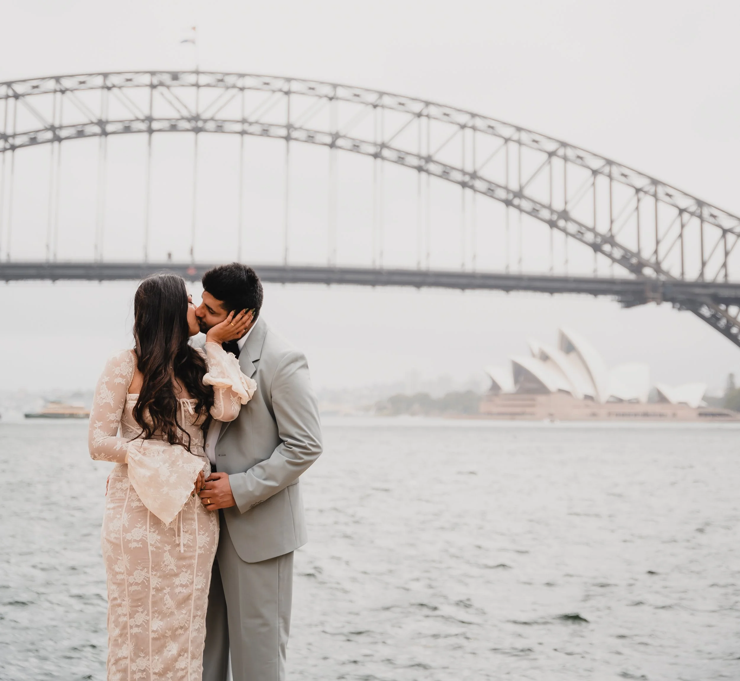 A couple is standing close and kissing by a river, with a large steel arch bridge in the background on a cloudy day.