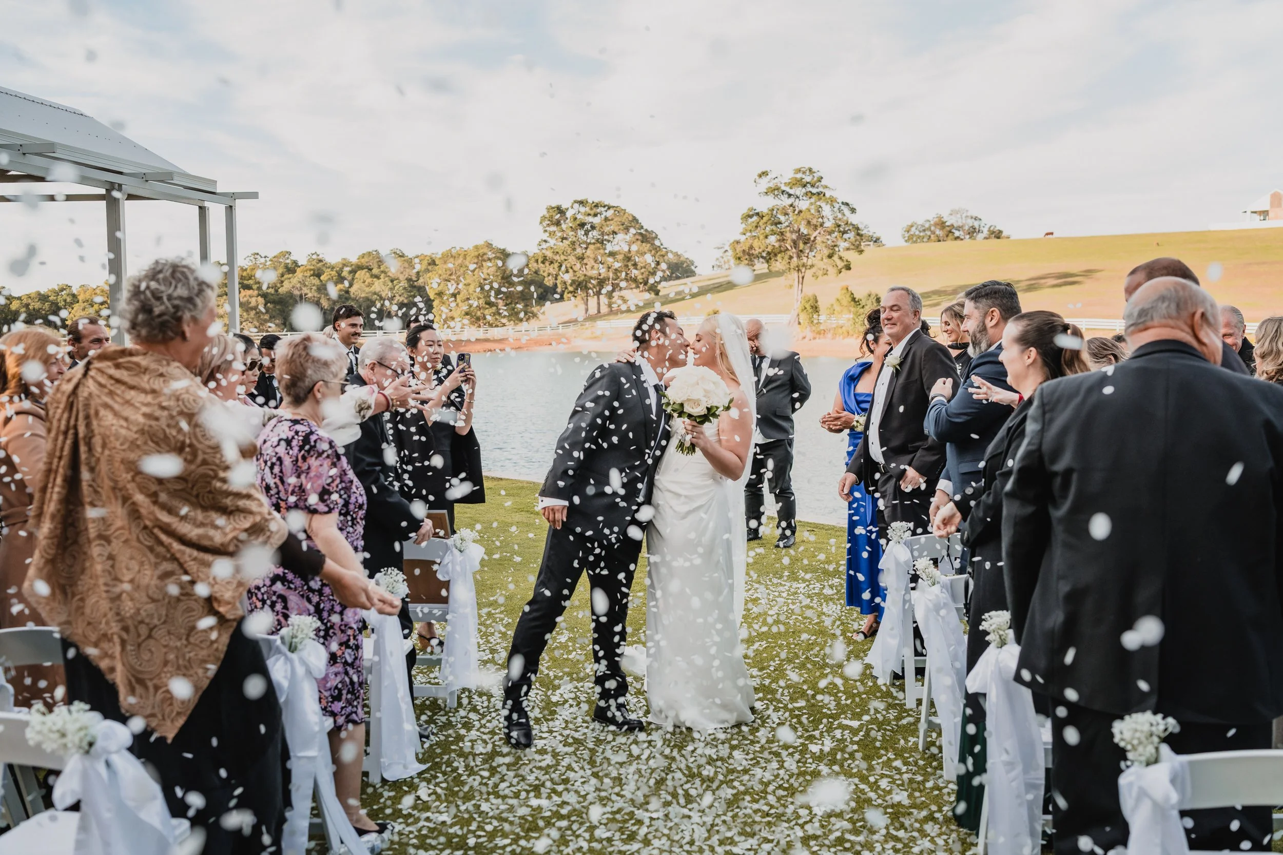 A couple at their outdoor wedding ceremony, kissing while surrounded by guests, with white rose petals falling from above on a grassy area near a lake and trees.