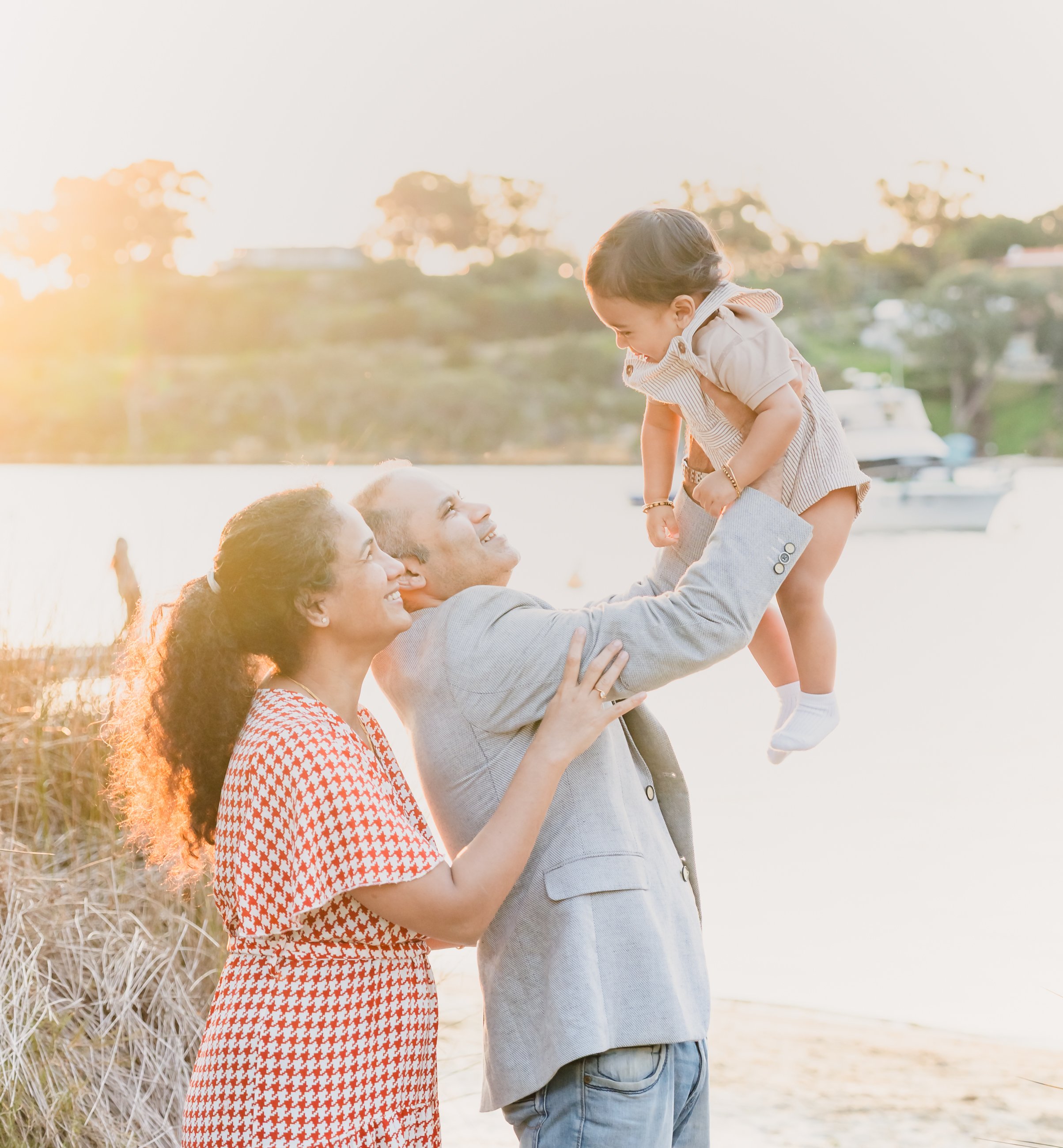 Family at the beach, a man lifts a young girl in the air as the sun sets in the background.
