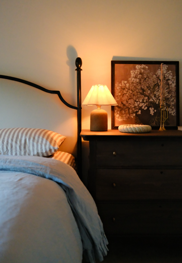 A cozy bedroom corner with a bed, a nightstand with a lit table lamp, a framed floral artwork, a decorative candle, and a white woven pillow.