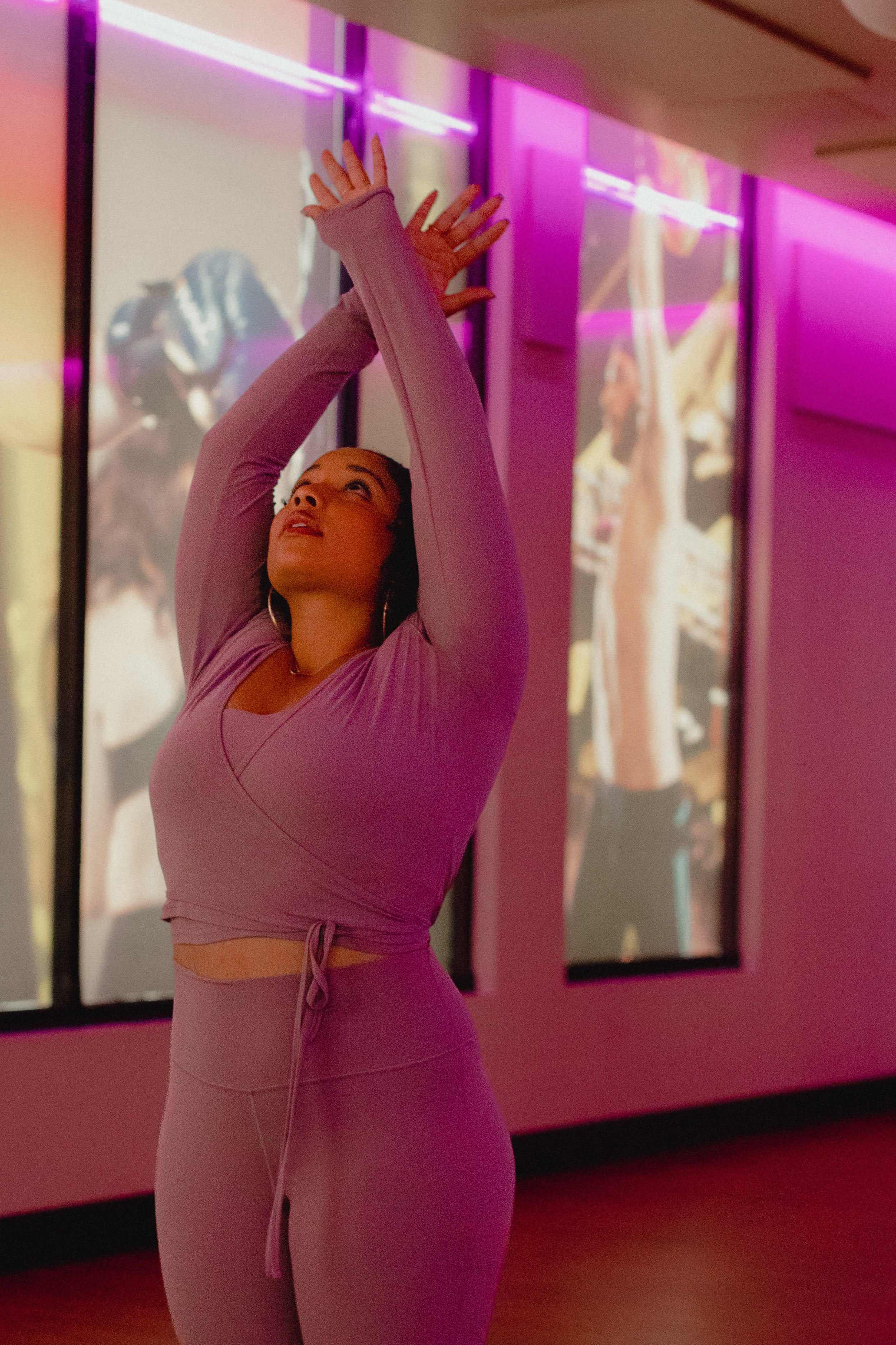 A woman in lavender athletic wear with long sleeves, standing in a room with pink and purple lighting and large digital displays in the background. She has her arms raised above her head, hands touching.