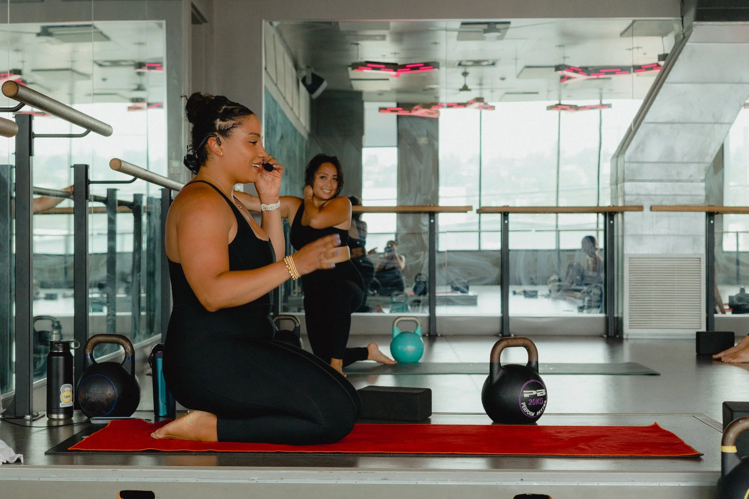 A woman practicing yoga in a gym, sitting on a red mat in a kneeling position with her legs folded under her and her hands relaxed on her thighs. She is wearing a black tank top and leggings, with her hair in braids, and a microphone headset. In the background, another woman is sitting on her heels, smiling, with fitness equipment and large windows visible.