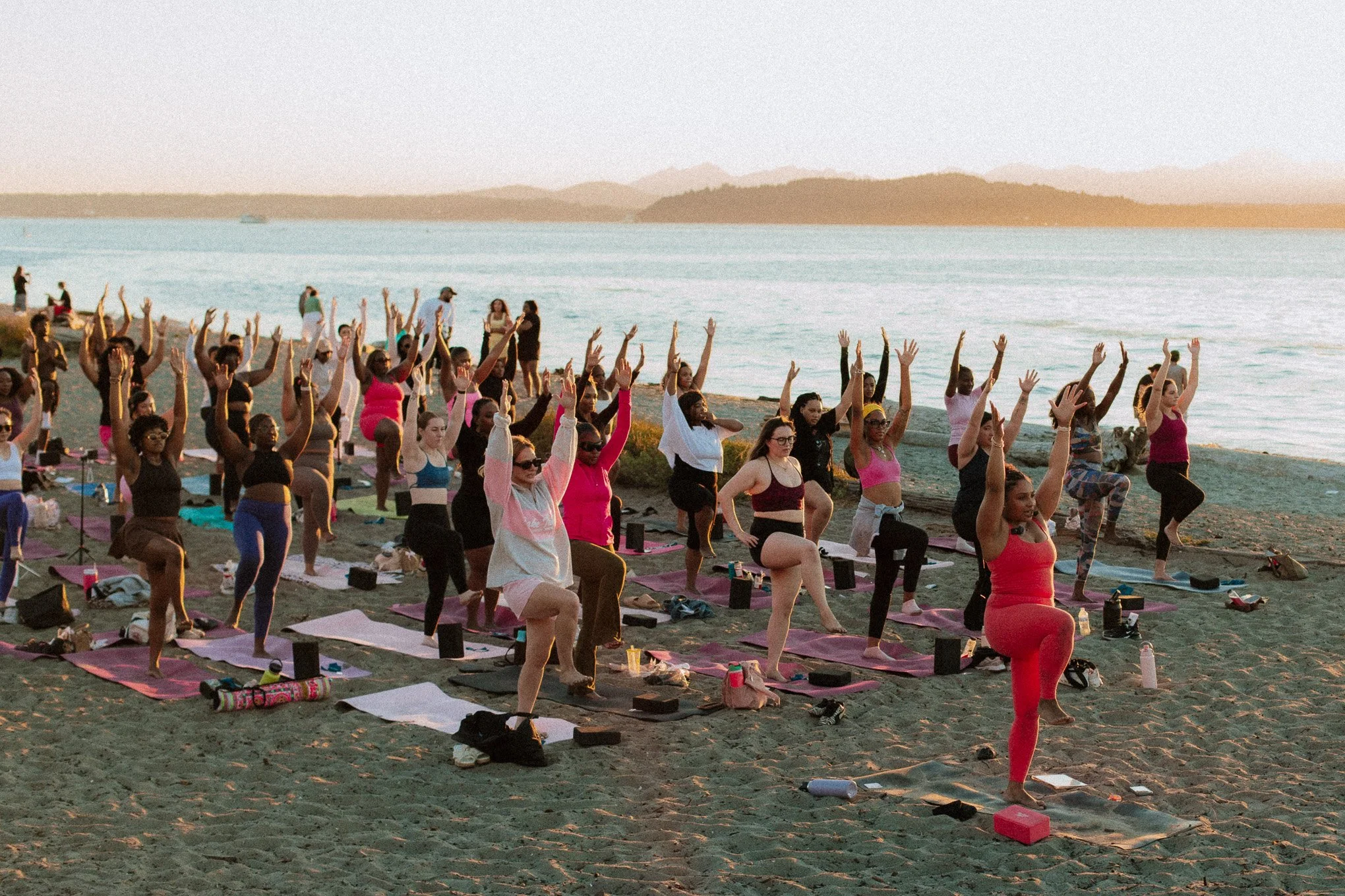 People participating in an outdoor yoga class on the beach during sunset, with the ocean and distant mountains in the background.