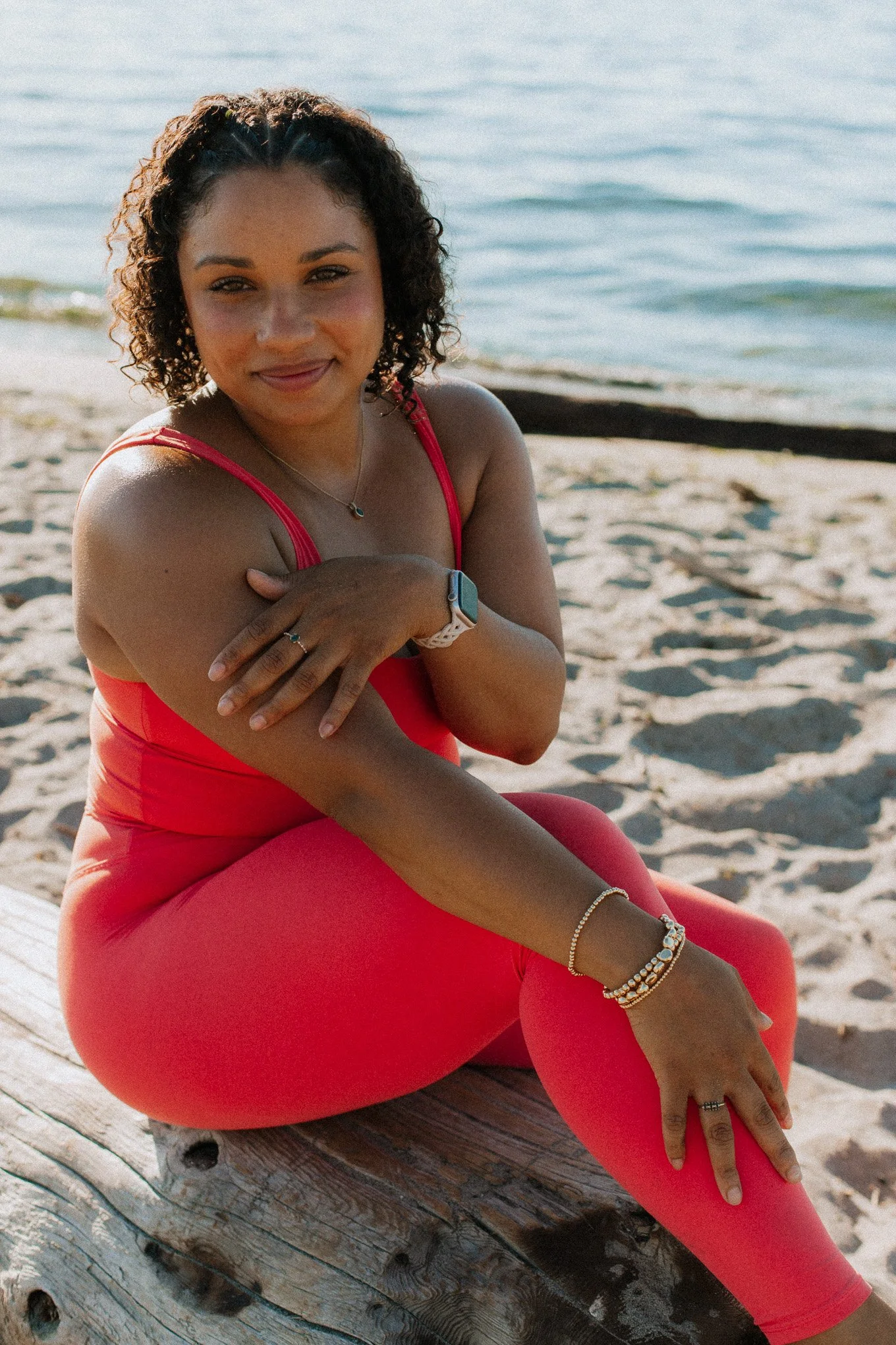 A woman with curly hair, wearing a pink top and matching leggings, sitting on a wooden log on the beach by the water, smiling at the camera.