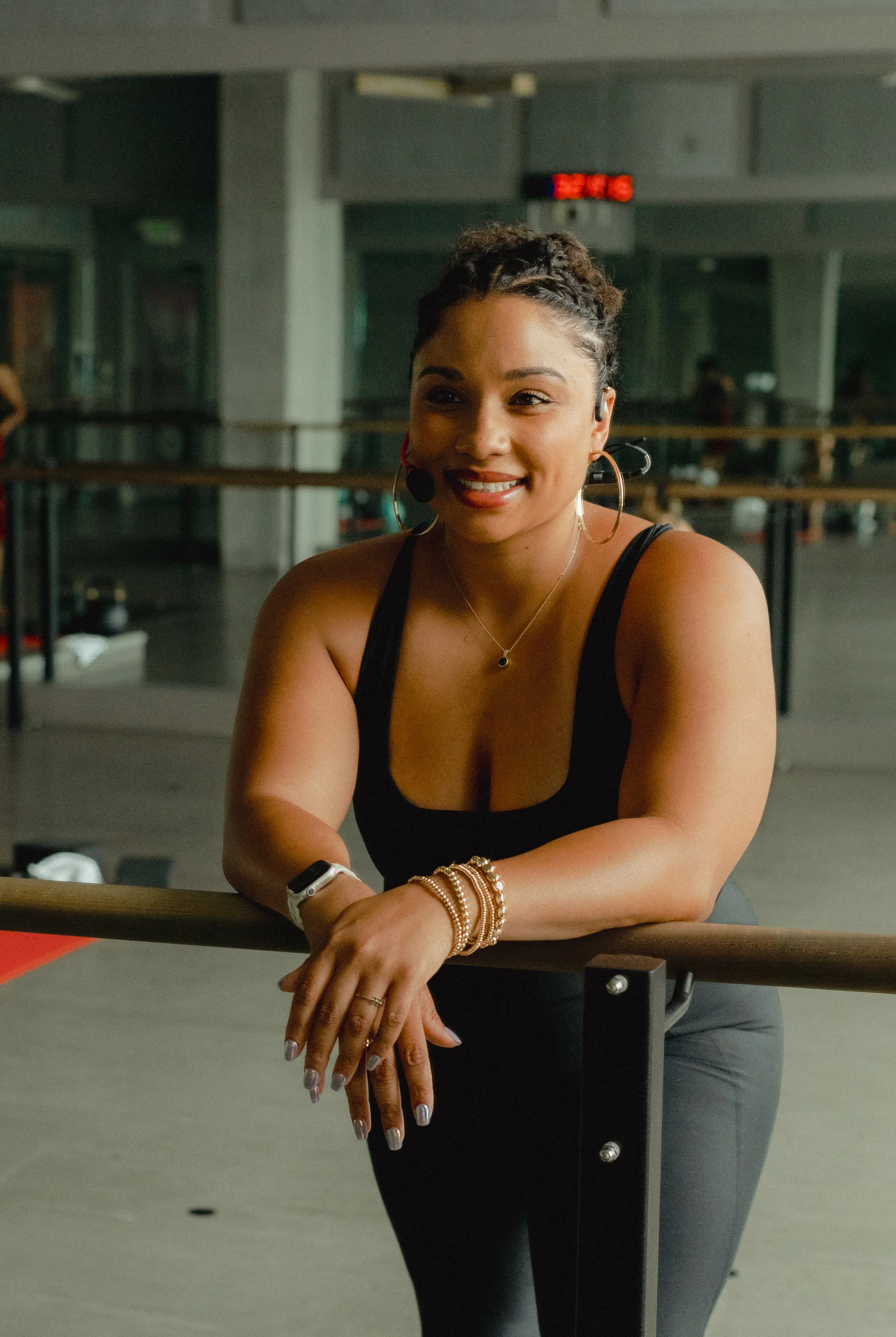 A woman with braided hair smiling and resting her hands on a ballet barre in a dance studio.