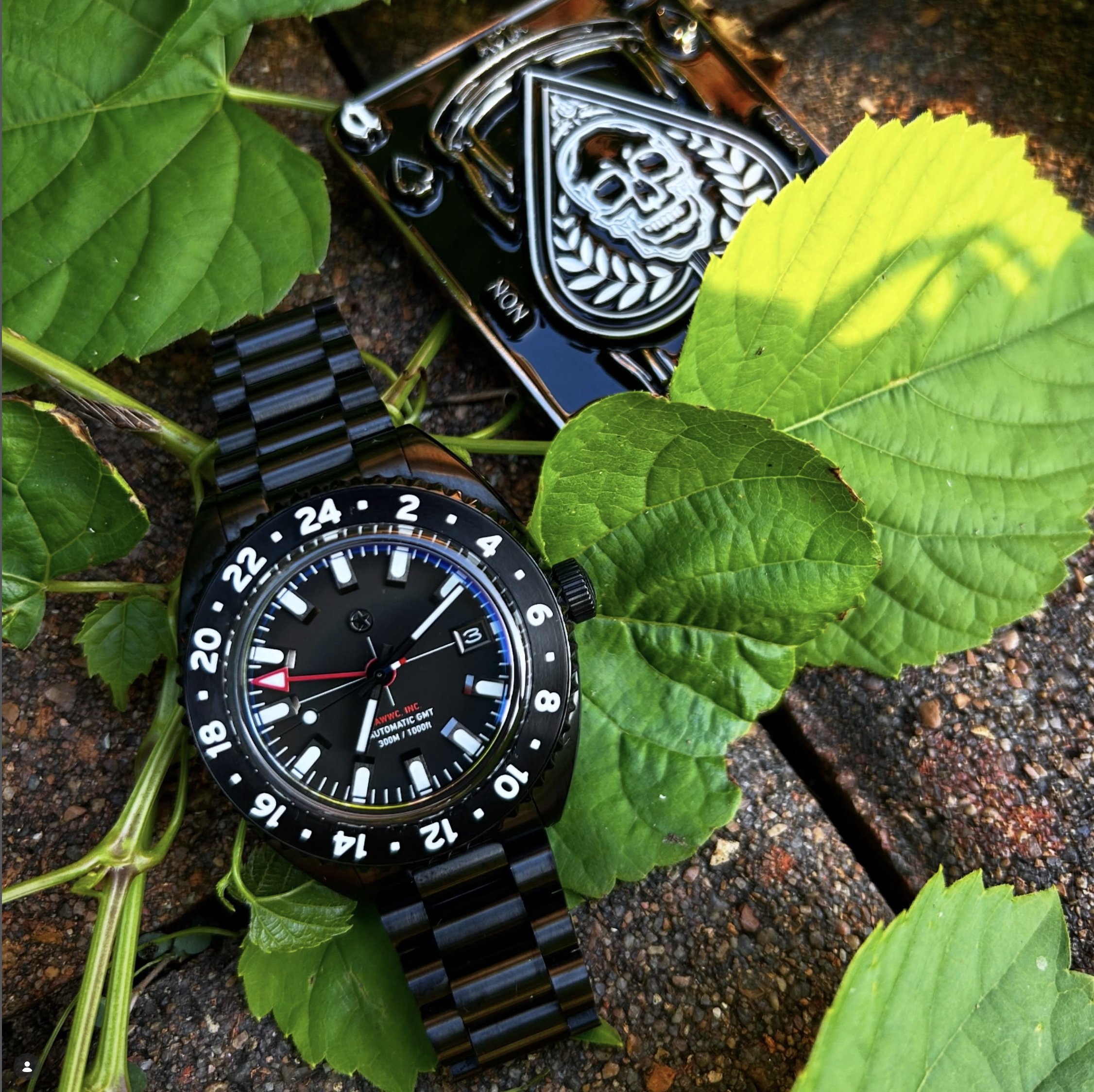 A wristwatch with a black band and a round face displaying a black dial, partially obscured by large green leaves on a rough, dark surface.