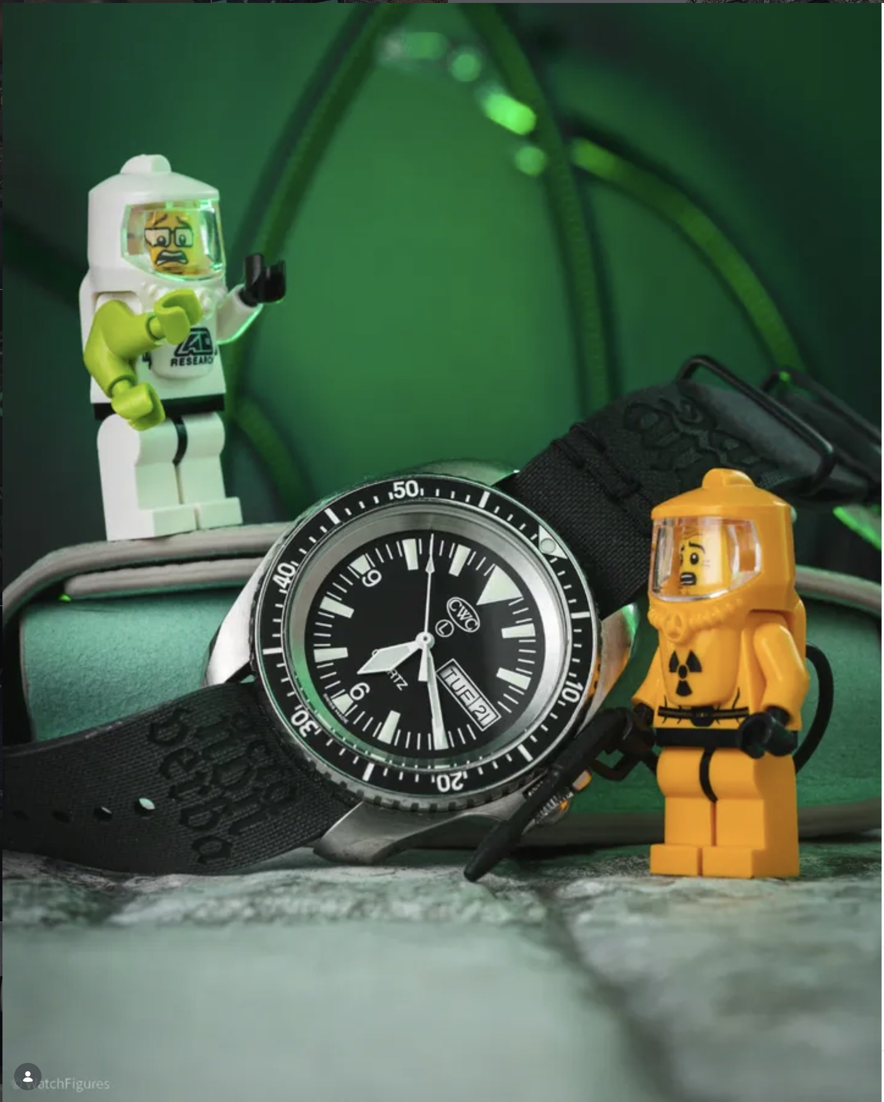 A LEGO yellow construction worker figure with a safety helmet and radioactive symbol on its chest standing next to a black strap watch with a silver frame. In the background, there are two small LEGO figures, one green and one yellow, with worried faces. The scene is set against a green and black background with cables.