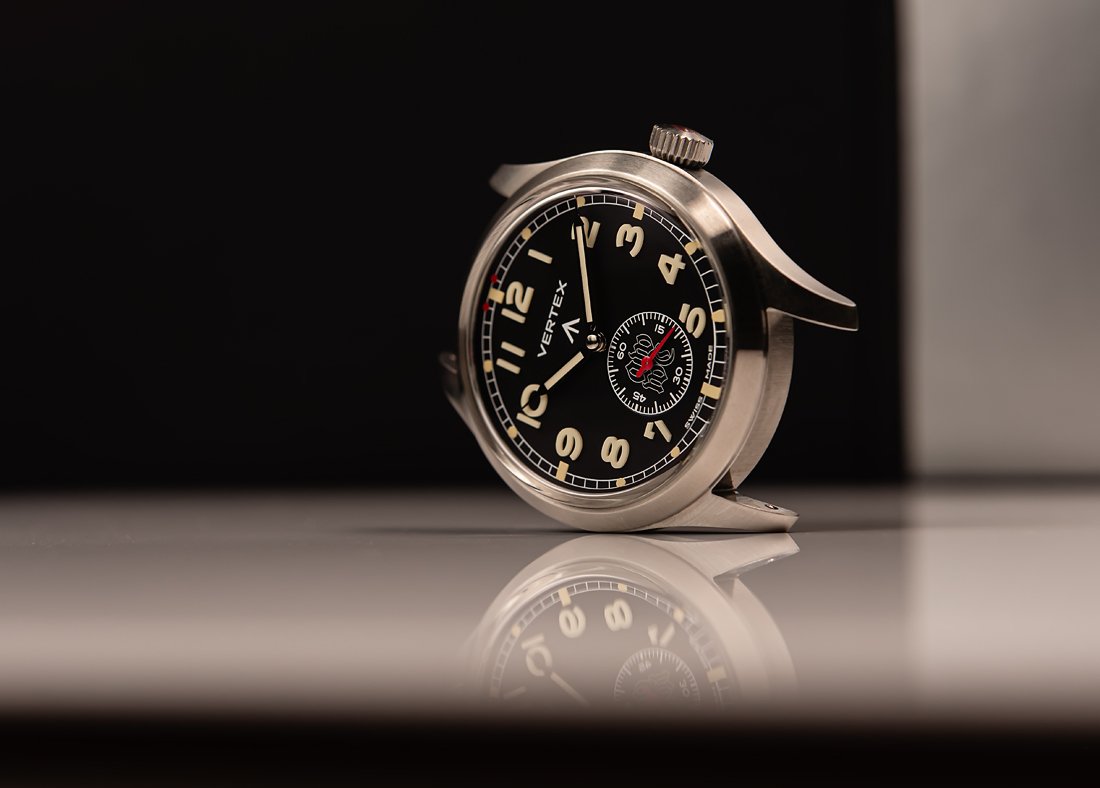 A close-up of a vintage-style wristwatch with a black dial, white numerals, and a small seconds sub-dial, resting on a reflective surface.