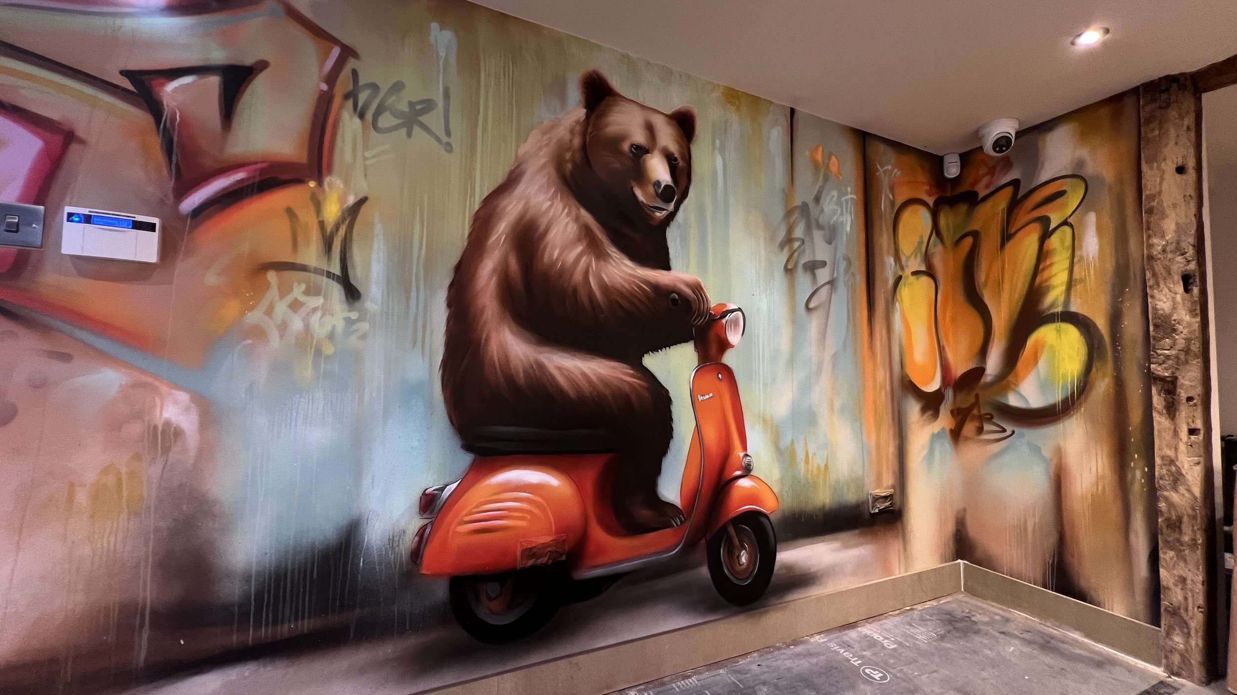 Graffiti wall mural of a brown bear riding an orange scooter, with colorful abstract graffiti background.