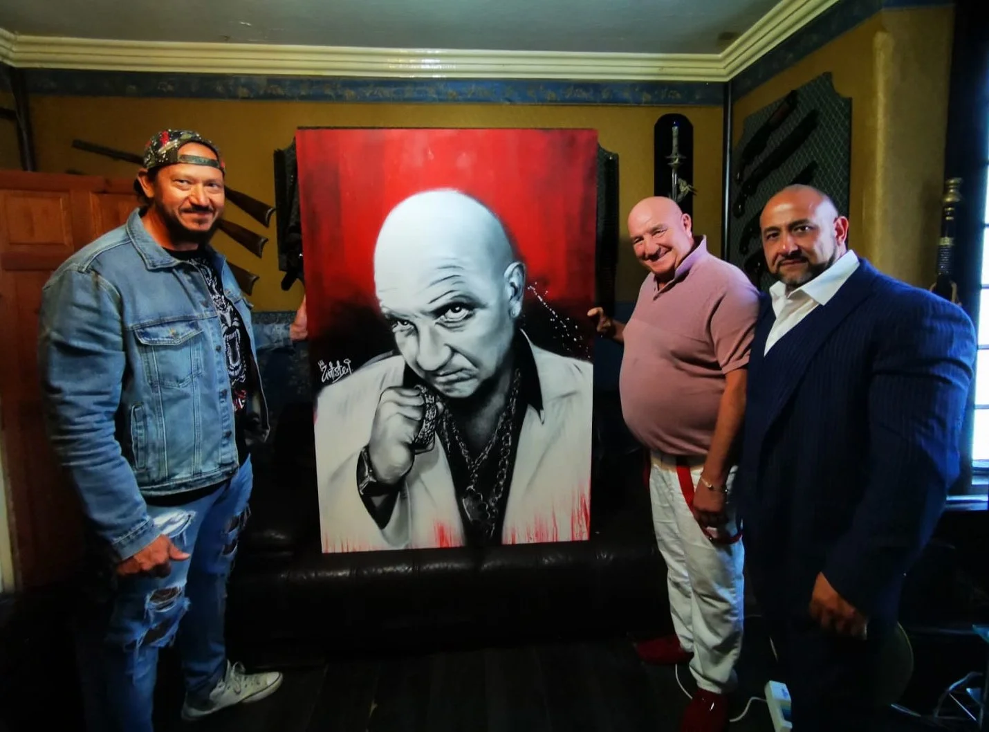 Three men standing on either side of a large painting of a bald man with a serious expression, wearing chains, with a red background. The men are smiling and posing for the photo.