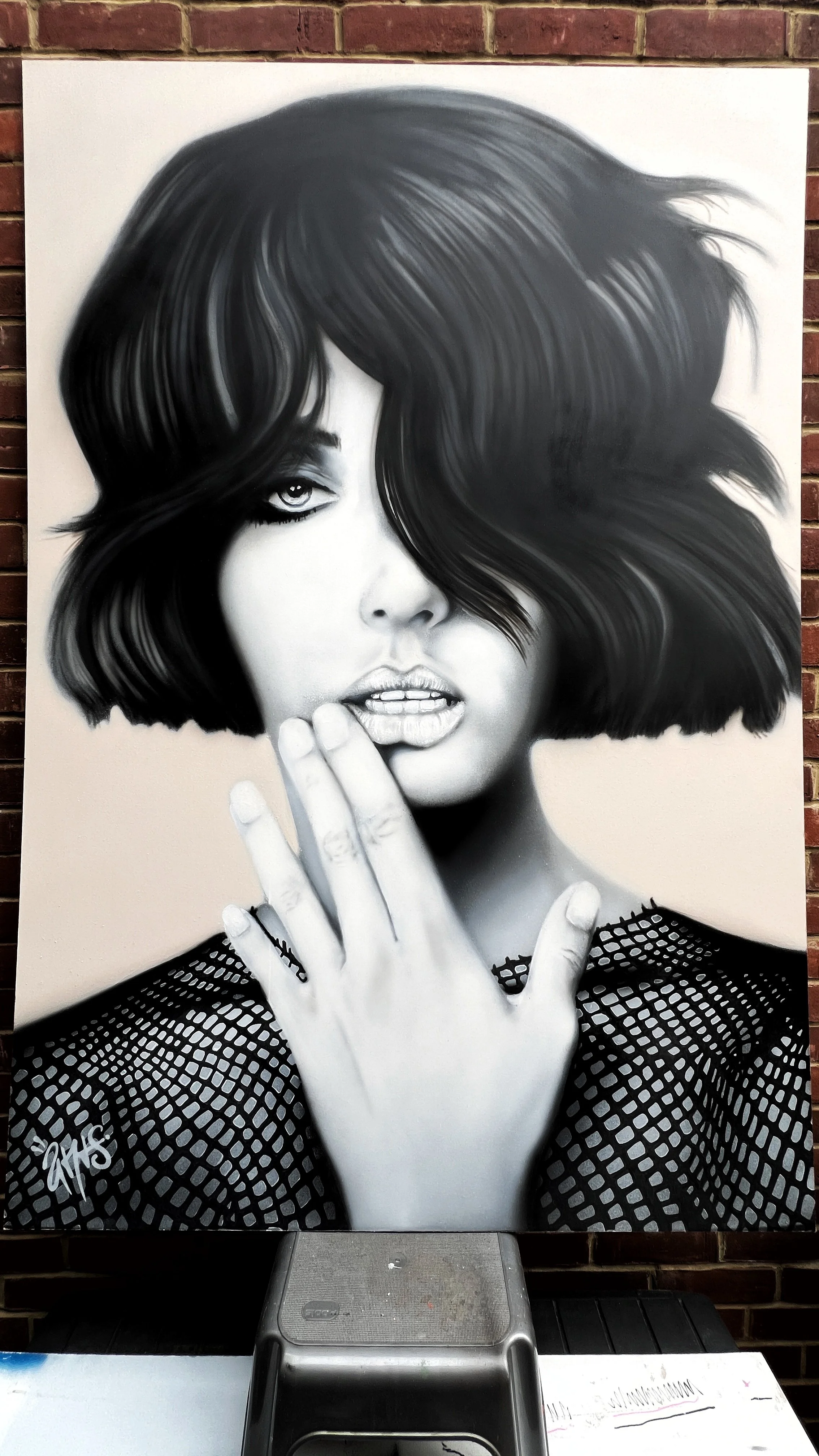 Black and white portrait of a woman with short, tousled hair covering one eye. She is touching her lips with her gloved hand, and the lower part of her face is partially covered by her hair. She is wearing a fishnet top.
