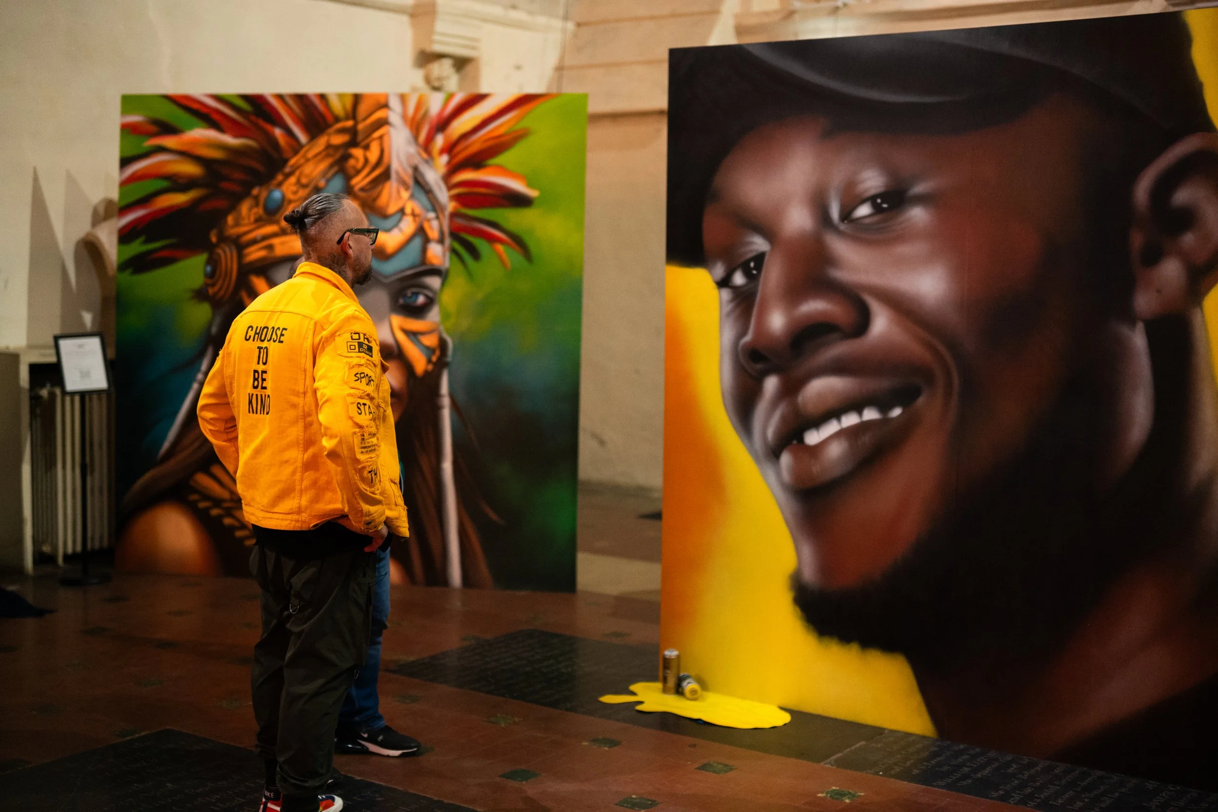 The artist Ants wearing a yellow jacket labeled "Choose to Be Kind" looking at large portrait prints of a girl with a joyful expression at an art exhibition.