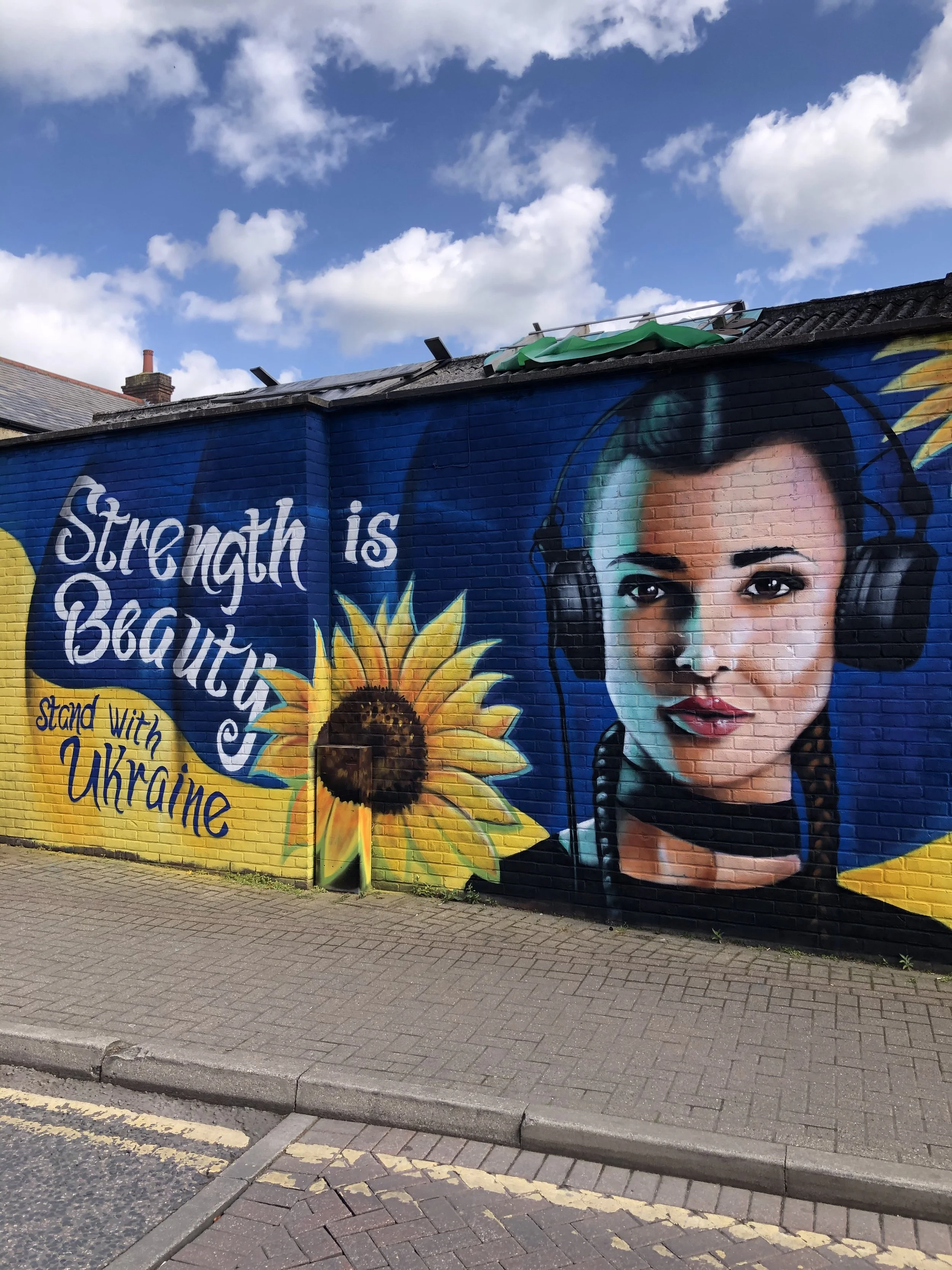 A mural on a brick wall depicting a woman with headphones, sunflowers, and text that reads 'Strength is Beautiful' and 'Stand with Ukraine'.