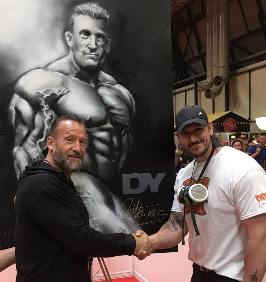 Two men shaking hands in front of a black and white portrait of a muscular man with the letters 'DY' at the bottom, in a busy indoor setting.