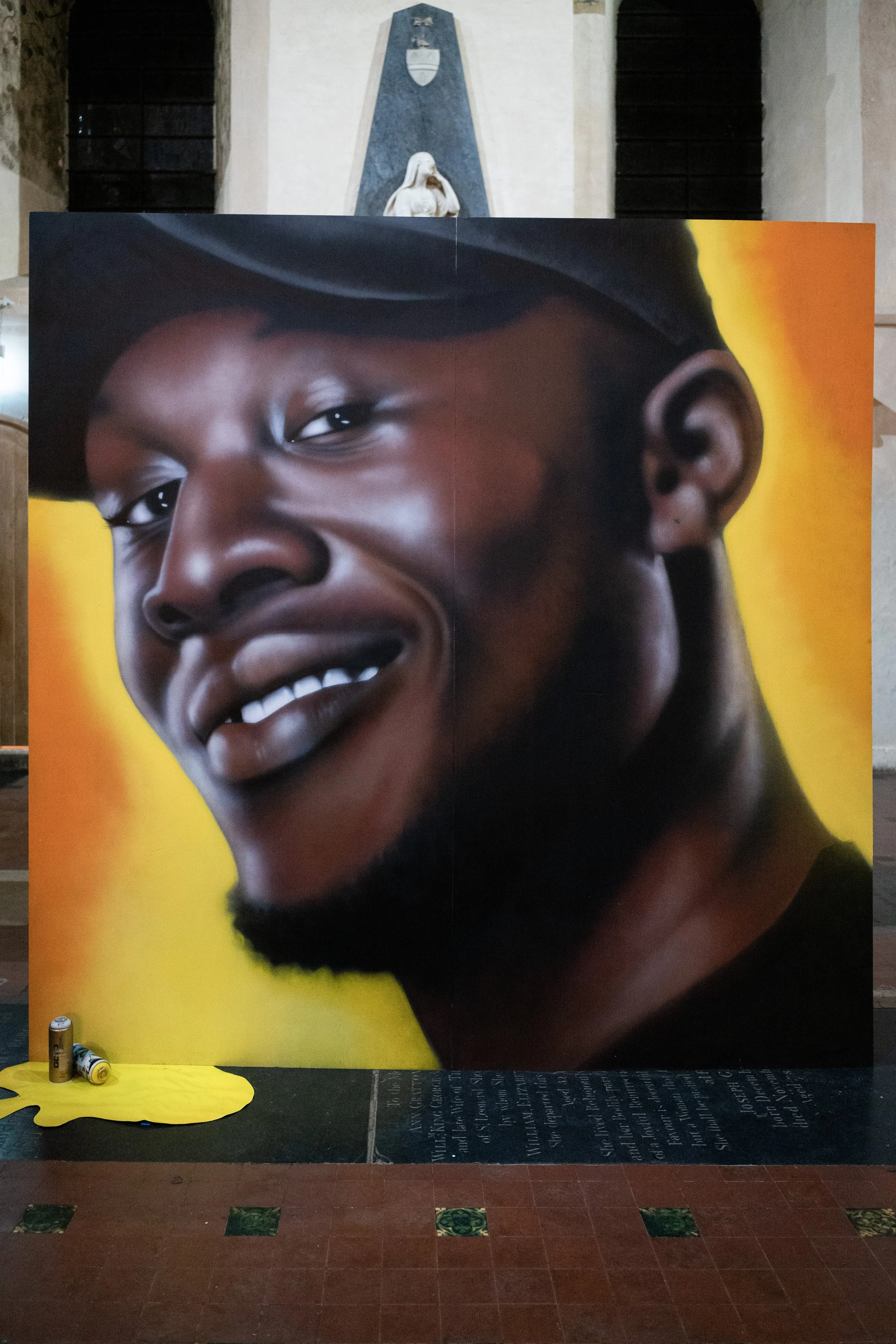 Large portrait of a smiling African American woman with short hair, set against a vibrant yellow background. The portrait is on a display board in a museum or gallery setting.