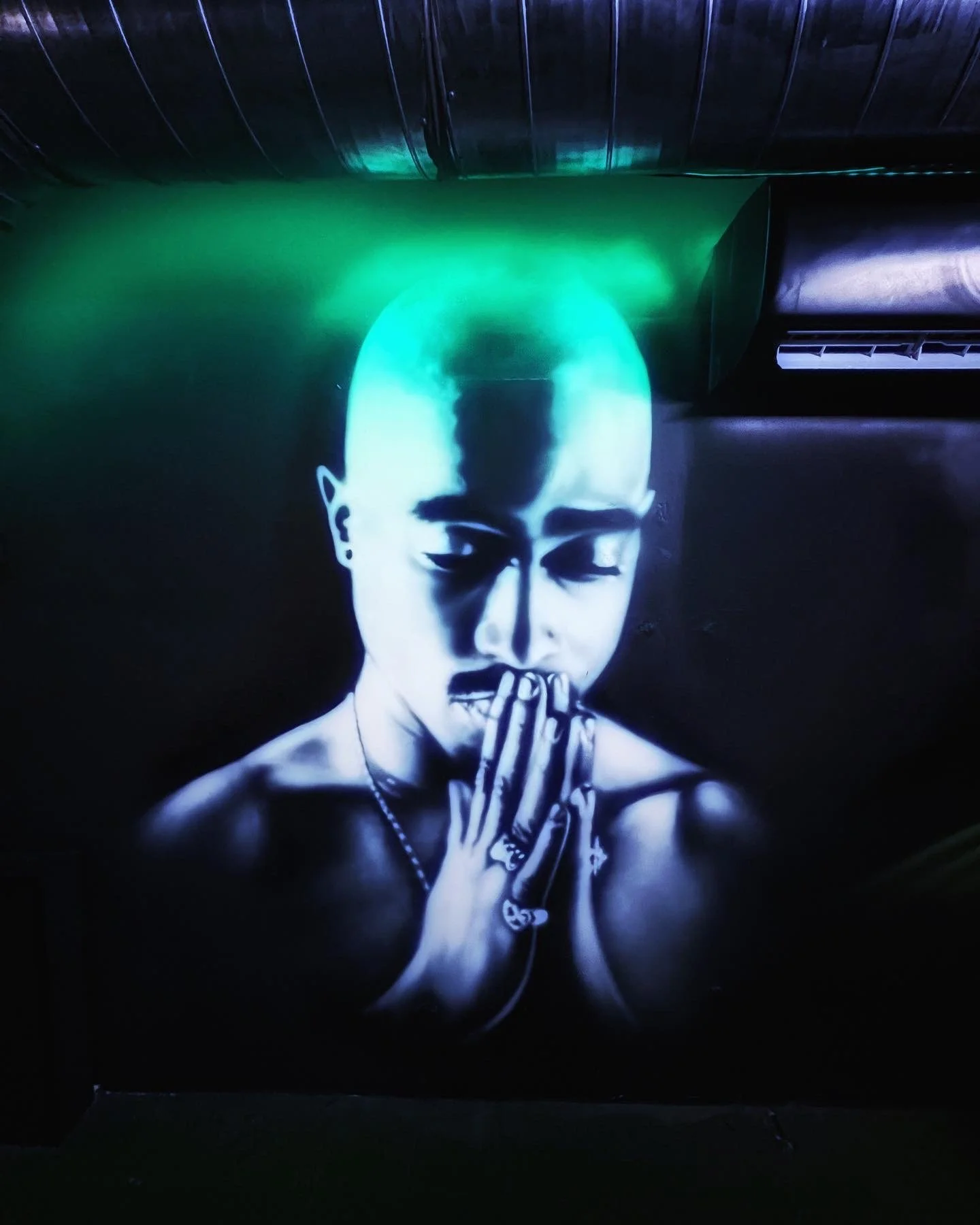 A black and white mural of a woman with shaved head, eyes closed, eyes covered with her hands, wearing a necklace, with green and blue lighting effects on her head and shoulders.