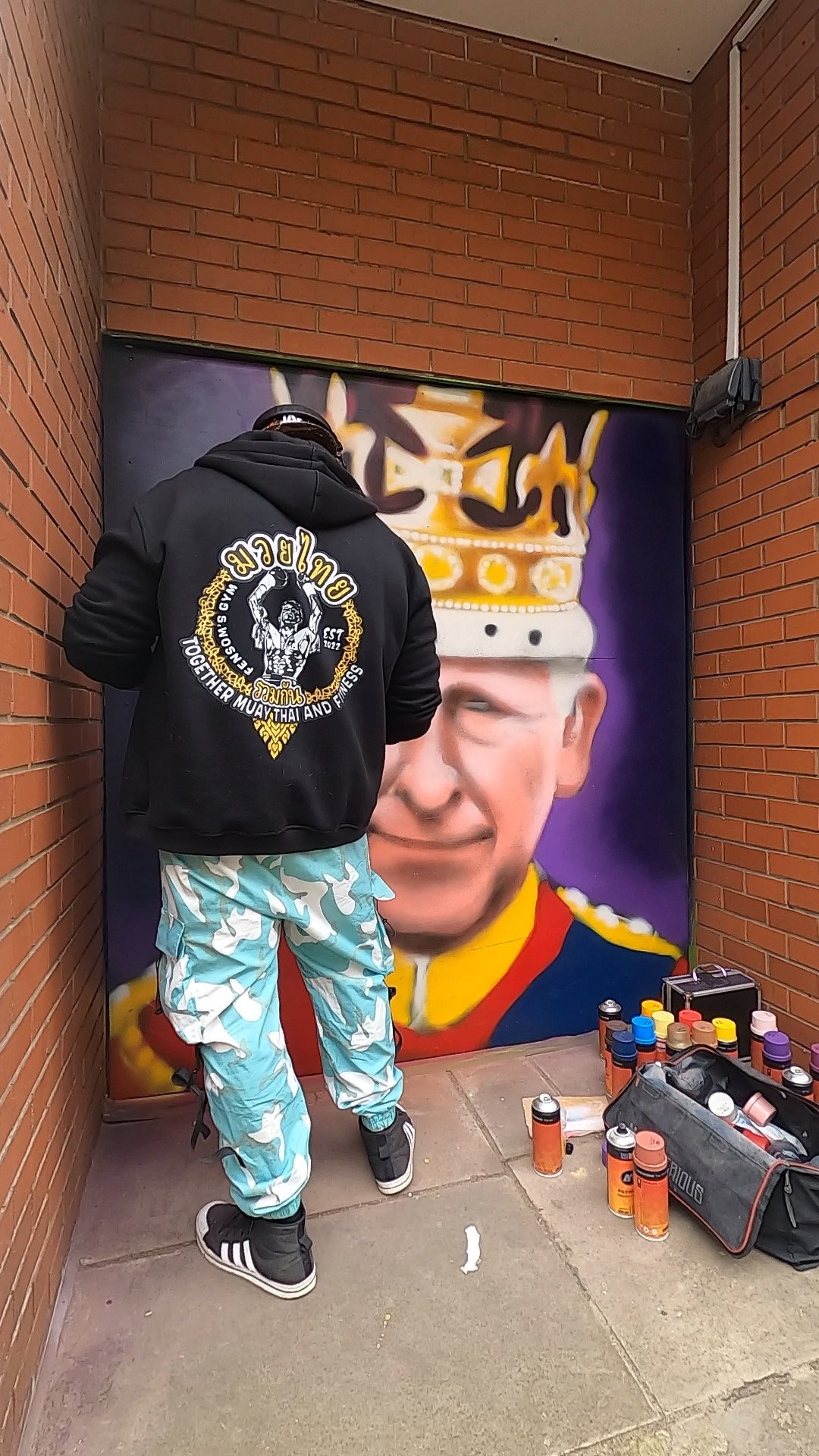 A person creating a mural of a young man wearing traditional Thai attire and a crown, with spray paint cans and supplies on the ground nearby.