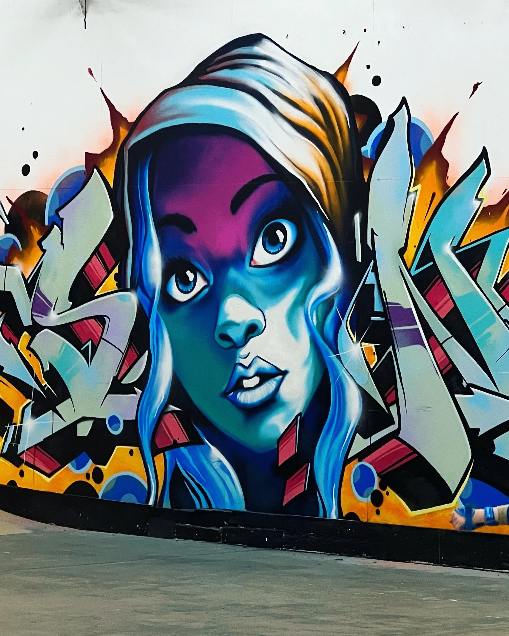 Street art mural of a woman with blue hair, expressive eyes, and winking, surrounded by abstract colorful shapes and patterns.