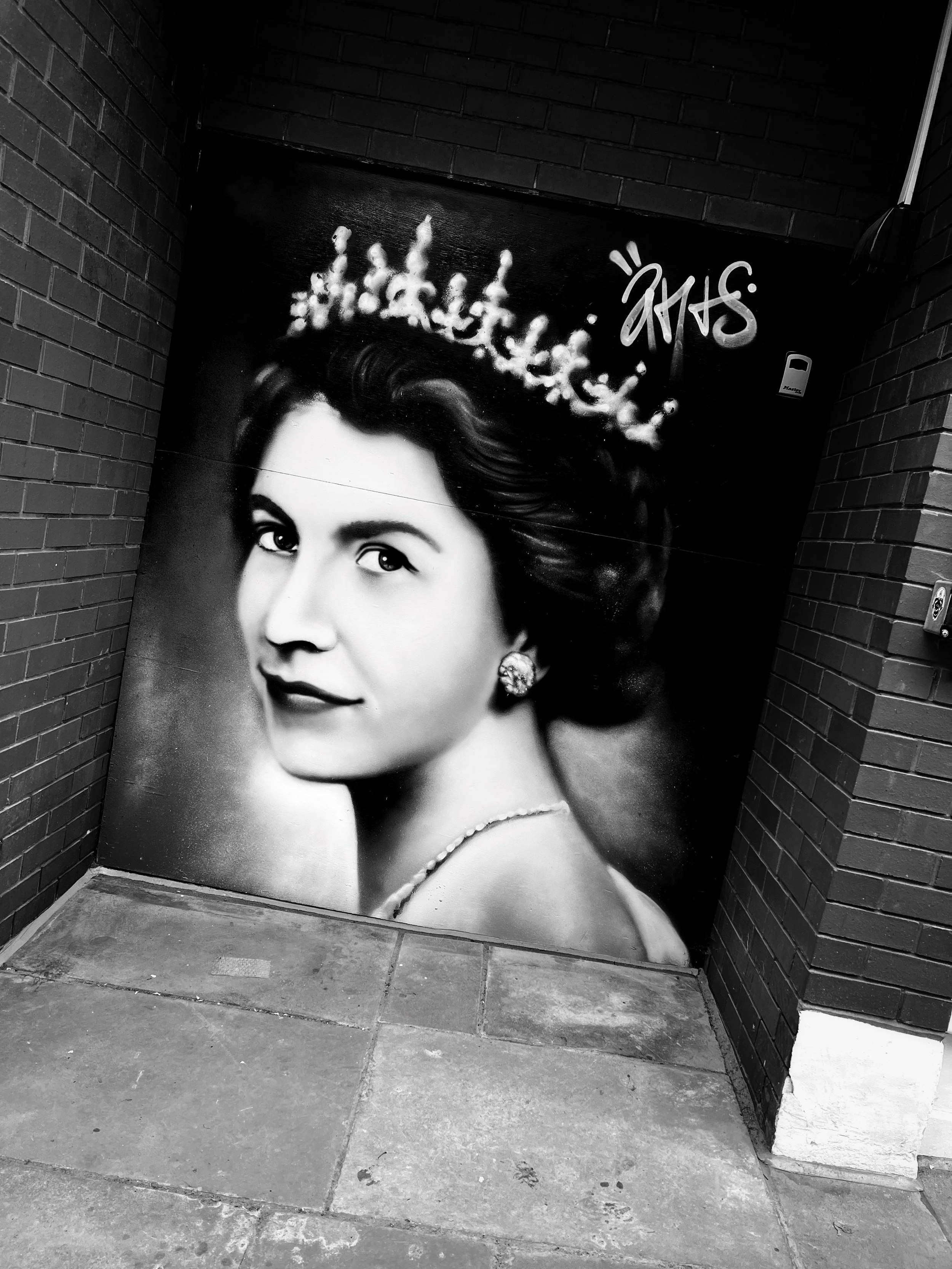 Black and white mural portrait of The Queen with styled hair, earrings, and a pearl necklace, set against a dark background with graffiti text.
