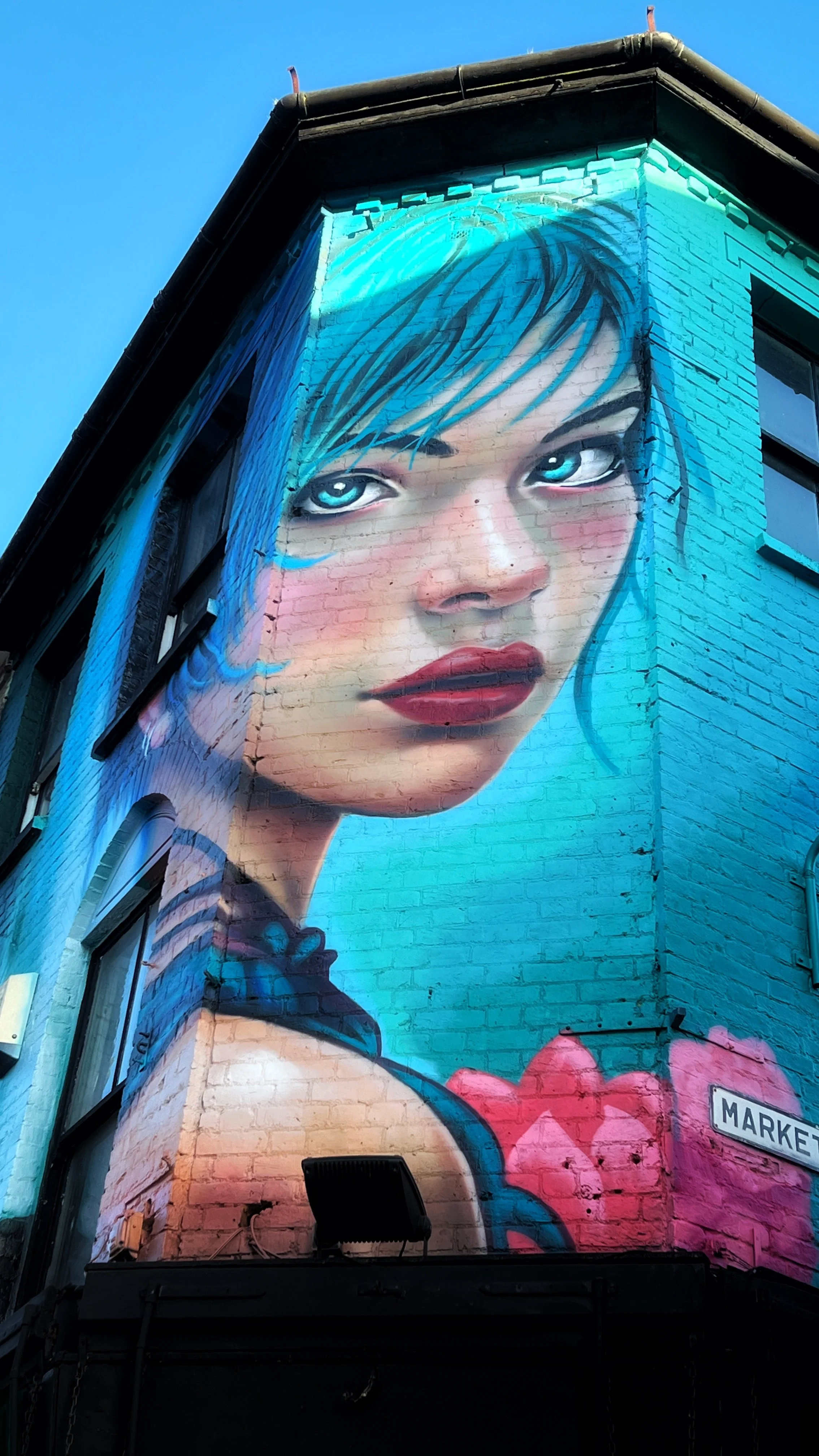 Large mural of a woman's face with blue eyes and blue hair, painted on the side of a brick building by artist Ants, titled "siren of the sea".