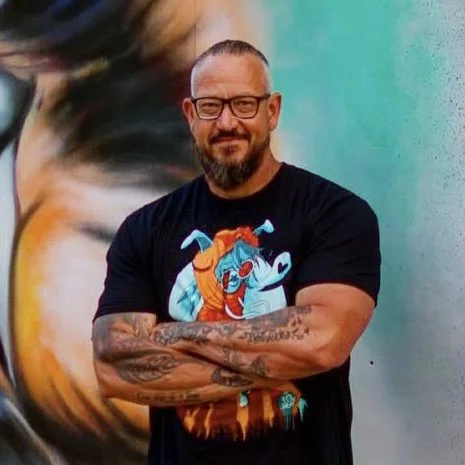 Man with glasses and a beard, wearing a black T-shirt with a colorful animated character, standing with crossed arms in front of a vibrant mural background.