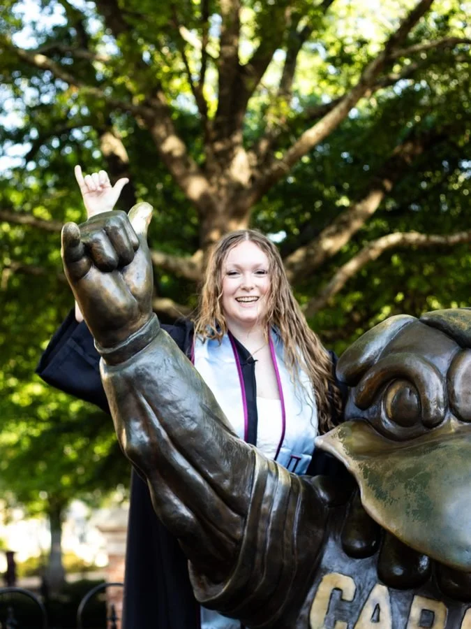 A young woman with long, curly hair in a graduation gown making a hand gesture while sitting on a large bronze sculpture of an eagle, with a tree in the background.
