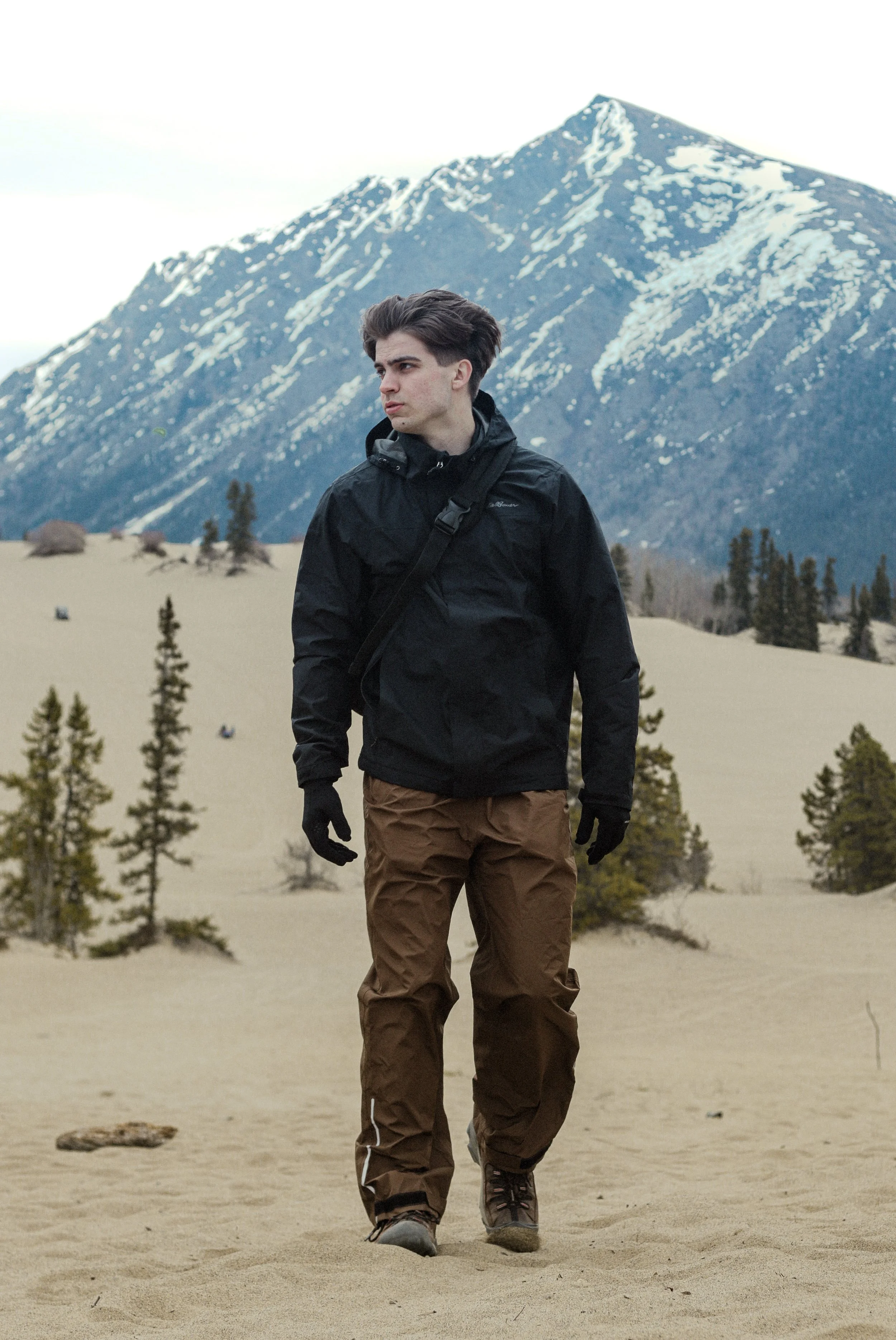 A young man walking in a sandy, desert landscape with a mountainous background. He is wearing a black jacket, brown pants, black gloves, and hiking boots.