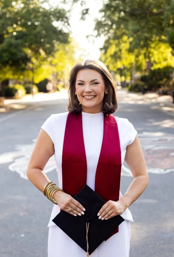 Young woman in a white outfit with a maroon stole holding a graduation cap, smiling outdoors on a sunny day.
