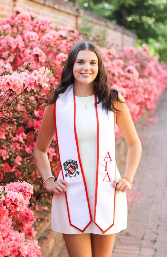 A young woman in a white dress standing on a sidewalk lined with pink azalea flowers, wearing a graduation stole with symbols and Greek letters.