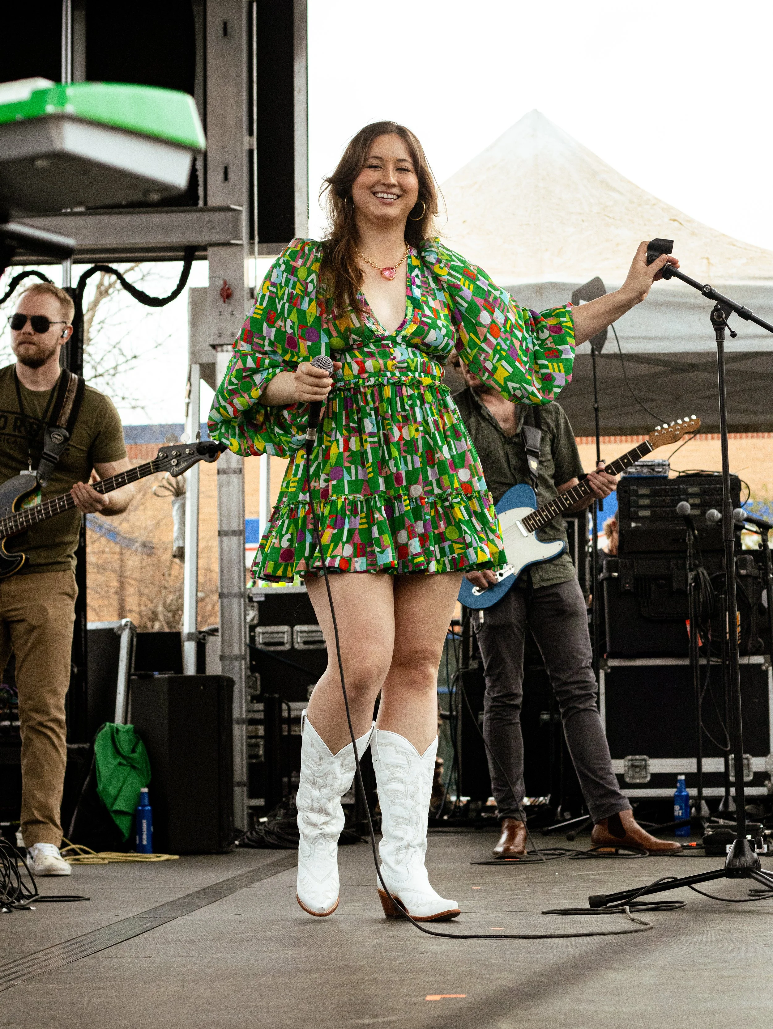 A woman wearing a colorful patterned dress and white cowboy boots performing on an outdoor stage with a microphone in her hand, smiling and facing forward, while band members play guitars behind her.