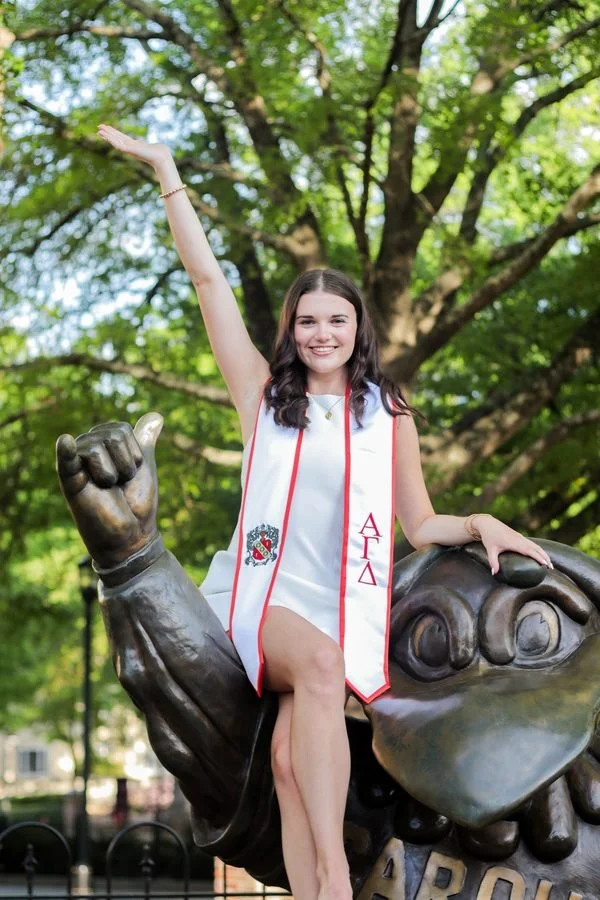 A young woman wearing a graduation stole, sitting on a statue of a large hand, raising her right arm in celebration, outdoors with green trees in the background.