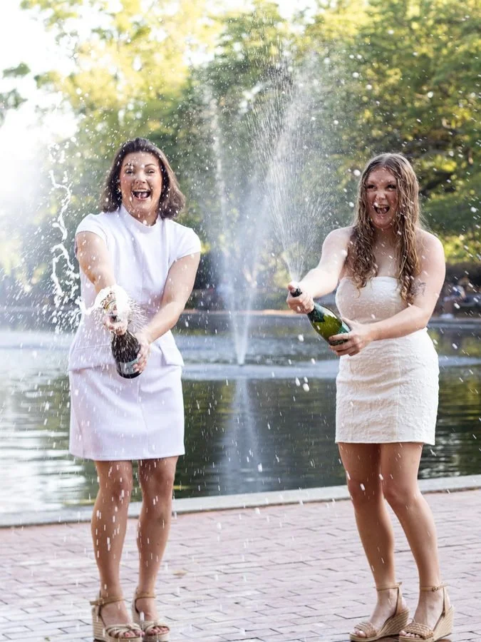 Two women celebrating outdoors near a body of water, wearing white dresses and champagne shoes, popping champagne bottles and spraying it in the air, smiling and having fun.