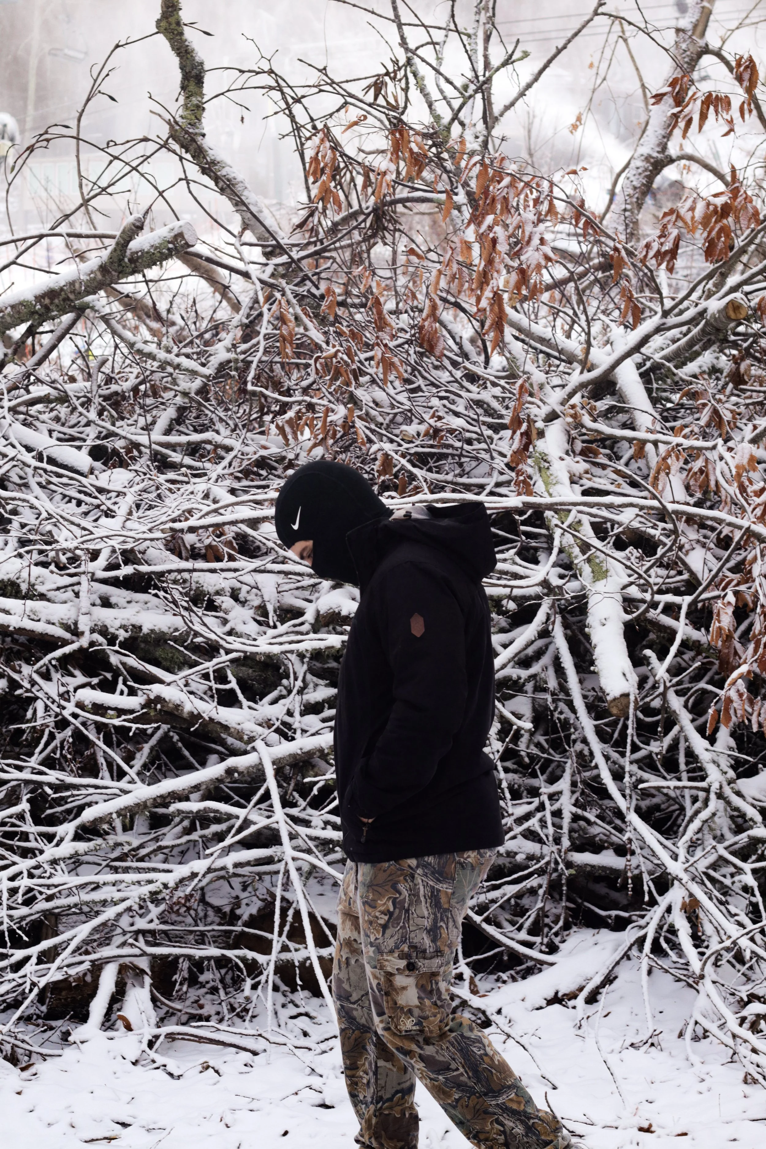 A person standing outdoors in front of a snow-covered fallen tree, wearing camouflage pants, a black jacket, and a black Nike beanie, with their head slightly bowed.