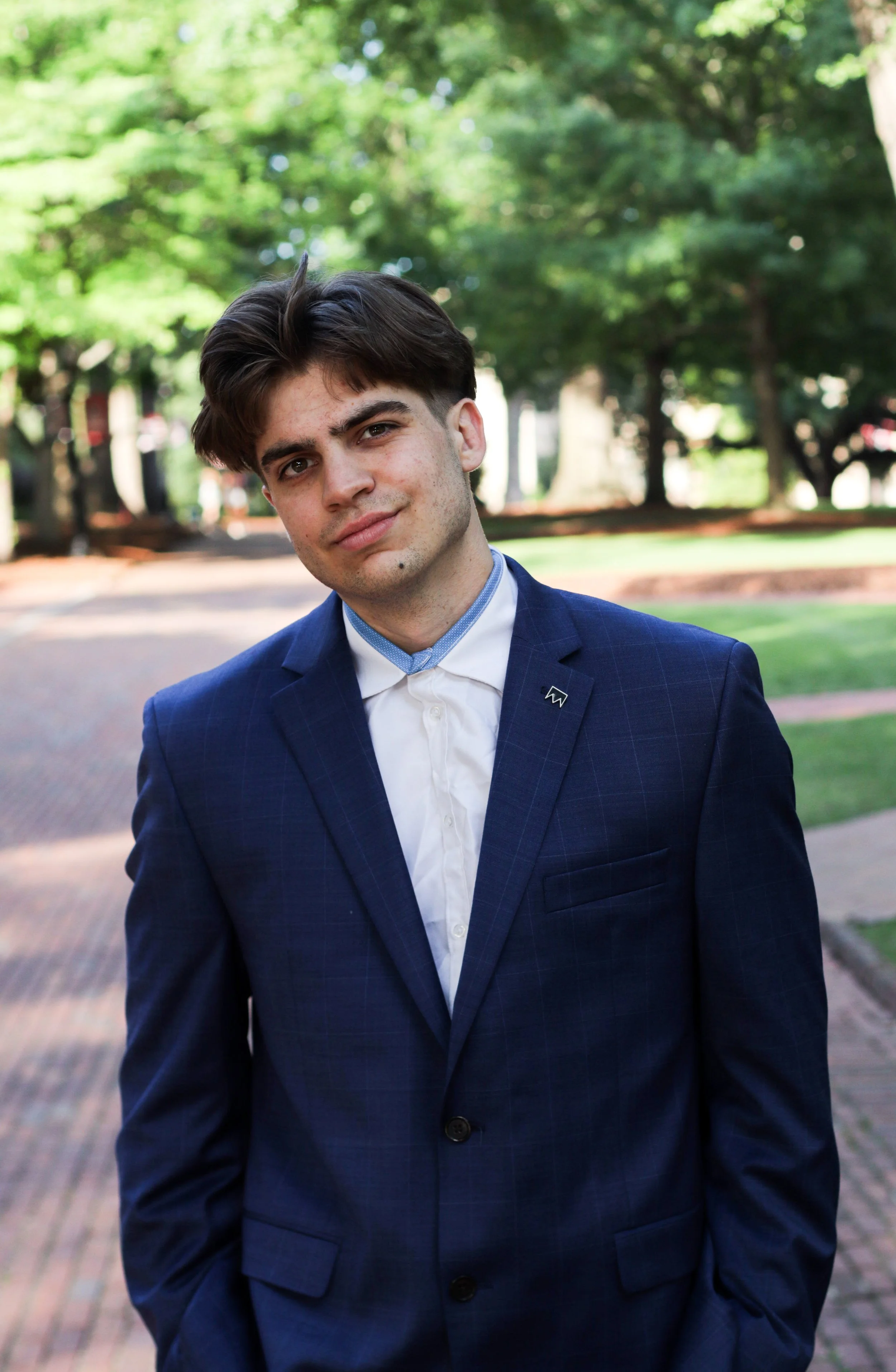 A young man in a blue suit and white shirt standing outdoors on a brick pathway with trees and greenery in the background.