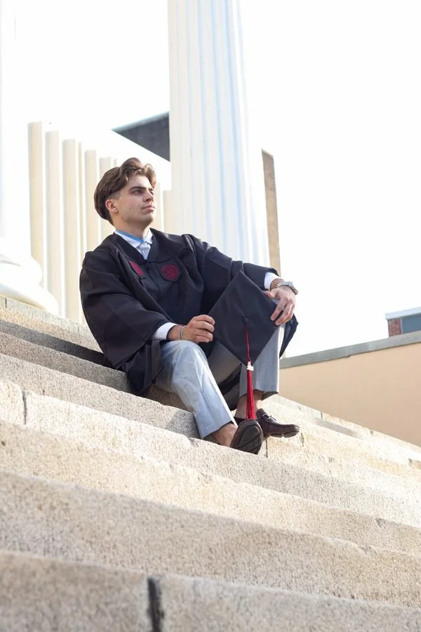 A young man in a graduation gown and cap sitting on outdoor steps, looking thoughtfully into the distance.