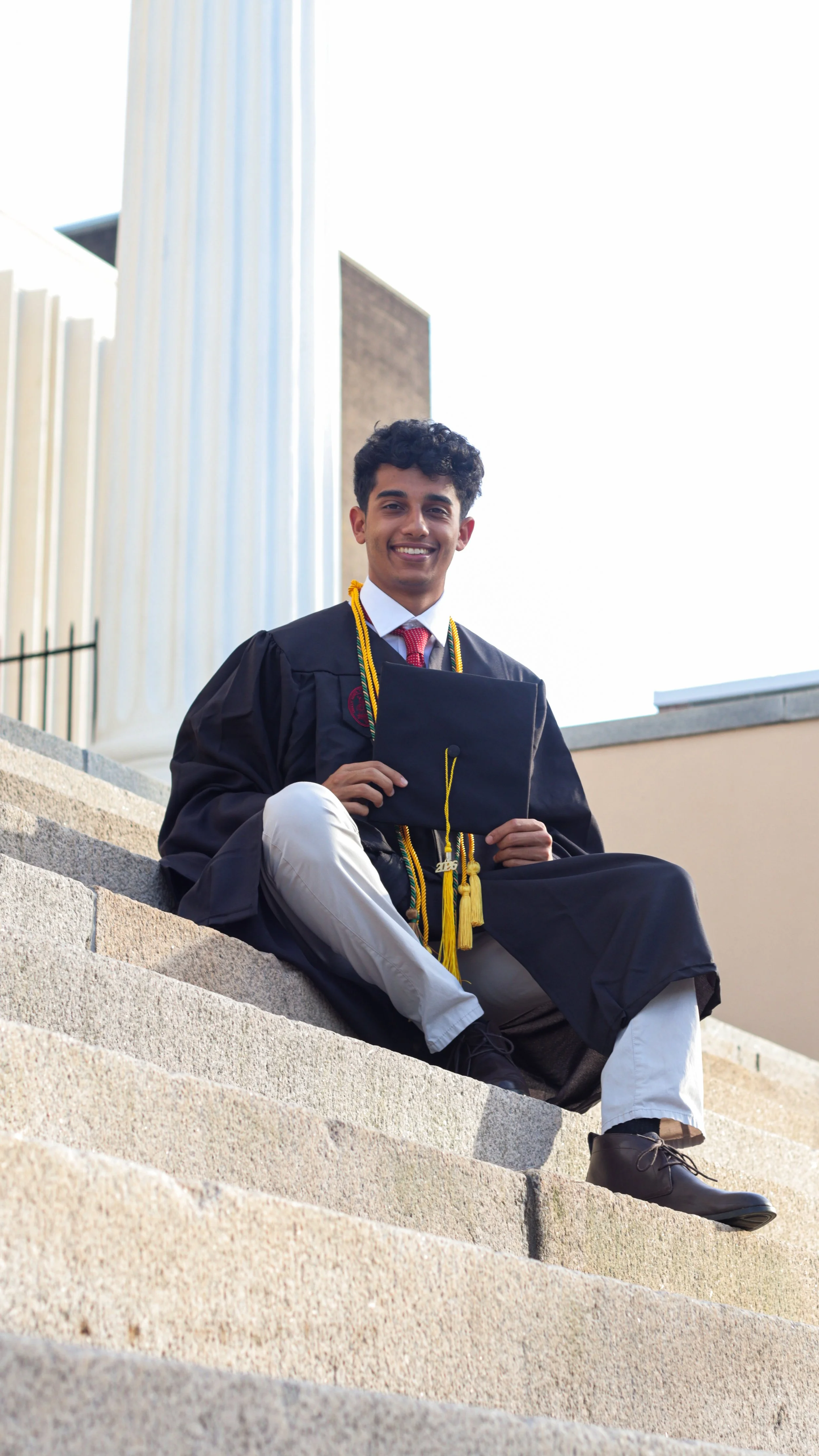 Young man in graduation gown and cap sitting on stone steps outdoors, smiling, holding cap, with a building and large white columns in the background.