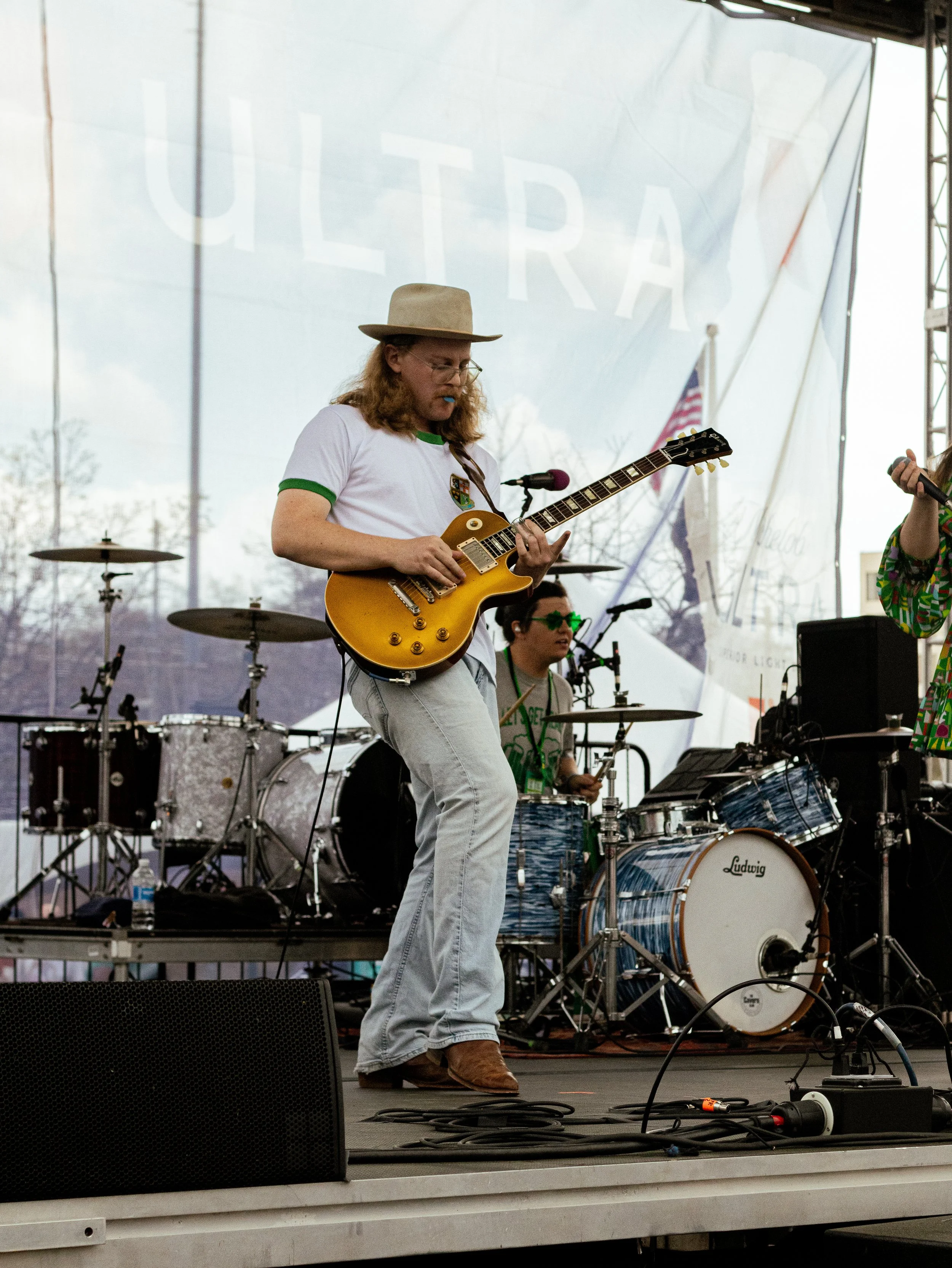 A person with long hair, glasses, wearing a beige hat and a white T-shirt with green trim, plays an electric guitar on stage. There are drums and other musical equipment behind them, and an American flag in the background. The stage is outdoors, like