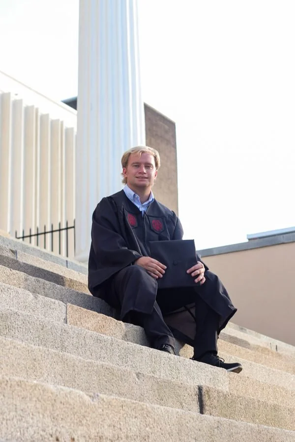 A young man in a graduation gown sitting on outdoor steps, holding a diploma, with a white column and building in the background.