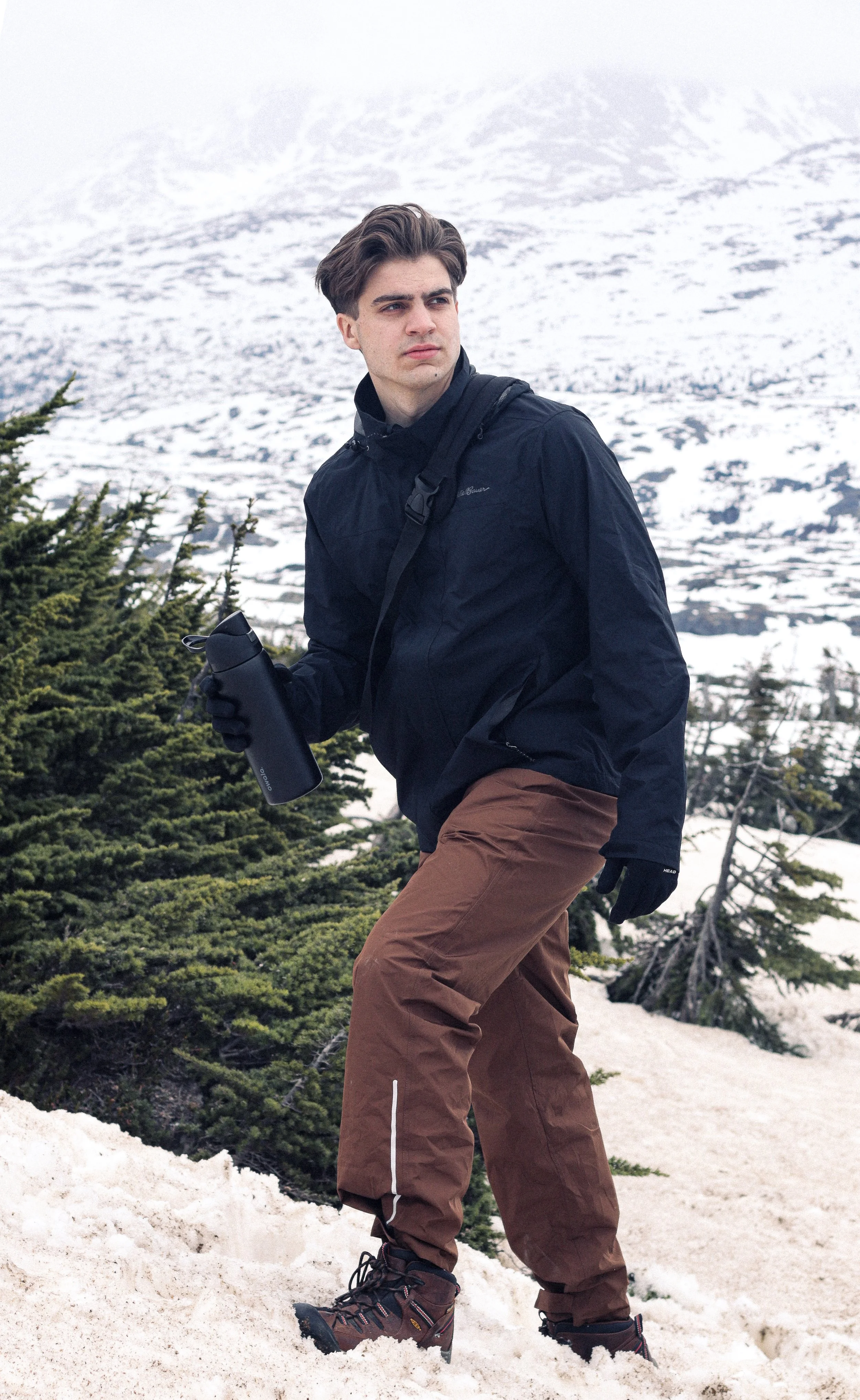 Young man in winter clothing and hiking boots, holding a water bottle, walking through snow-covered terrain with evergreen trees and snow-covered mountains in background.