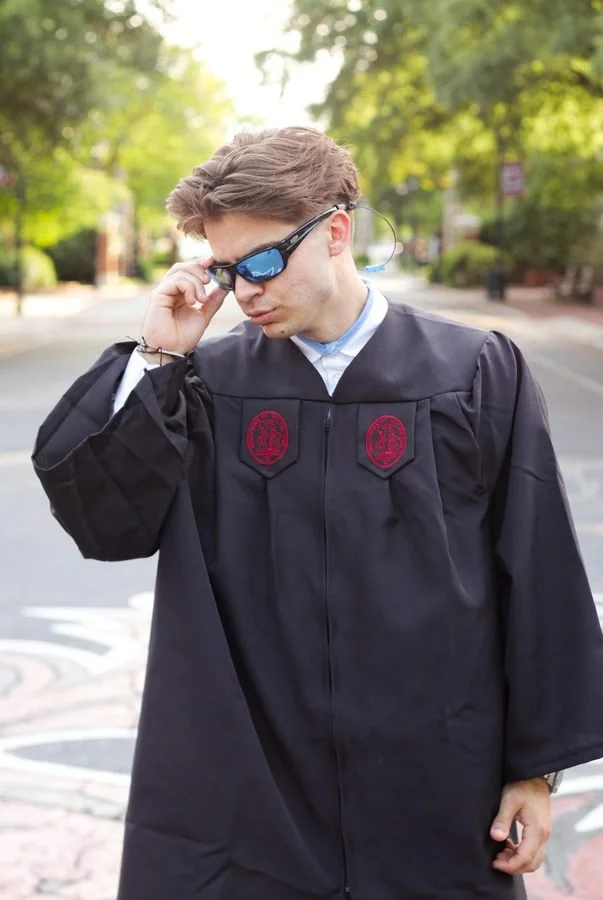 Young man in graduation gown wearing sunglasses on a sunny street.