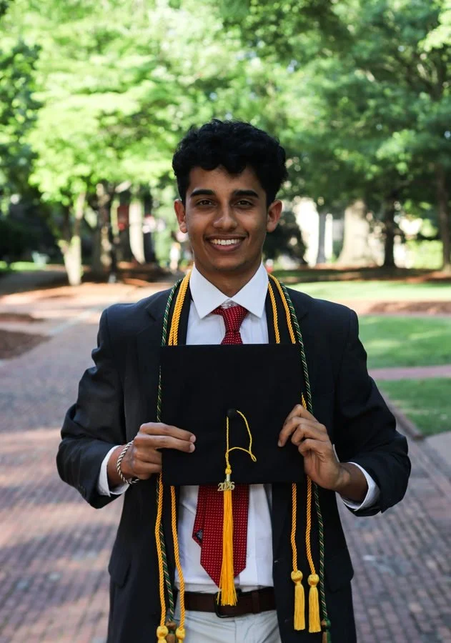 Young man in graduation gown and cap holding diploma outdoors in a park with green trees in the background.
