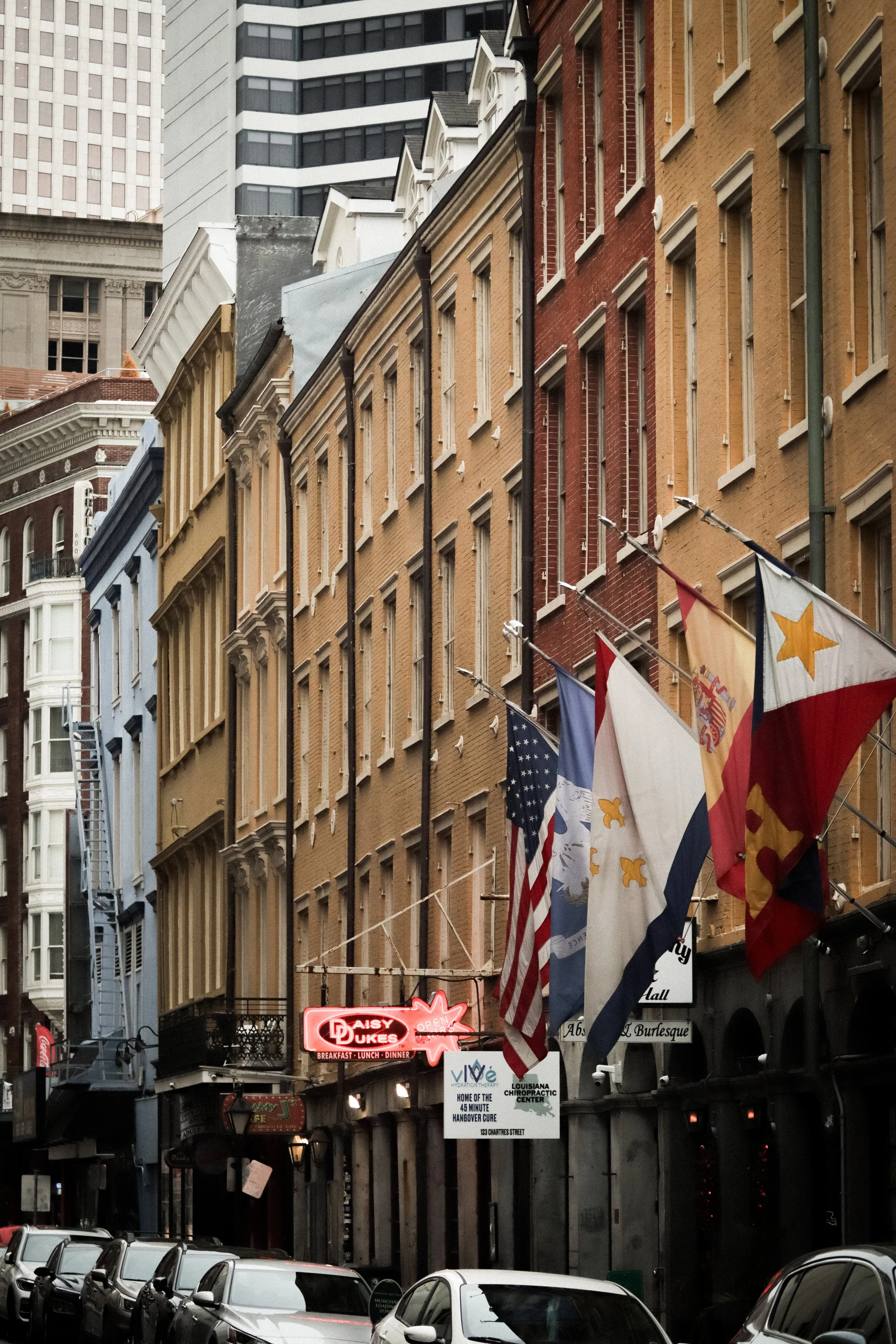 Downtown street scene with a row of historic brick and stone buildings featuring decorative facades and multiple windows, with flags hanging in front. Cars are parked along the street, and neon signs are visible at street level, including a red sign reading 'Daisy Dukes'.