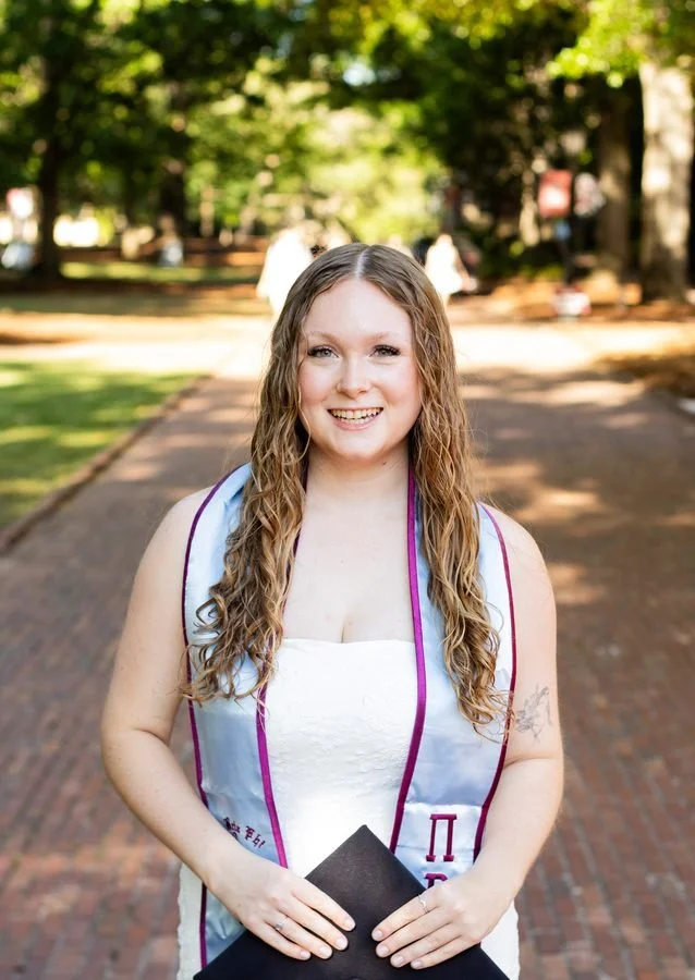 A young woman with long, curly hair smiling and holding a diploma outside on a sunny day, walking on a brick pathway in a park-like setting.