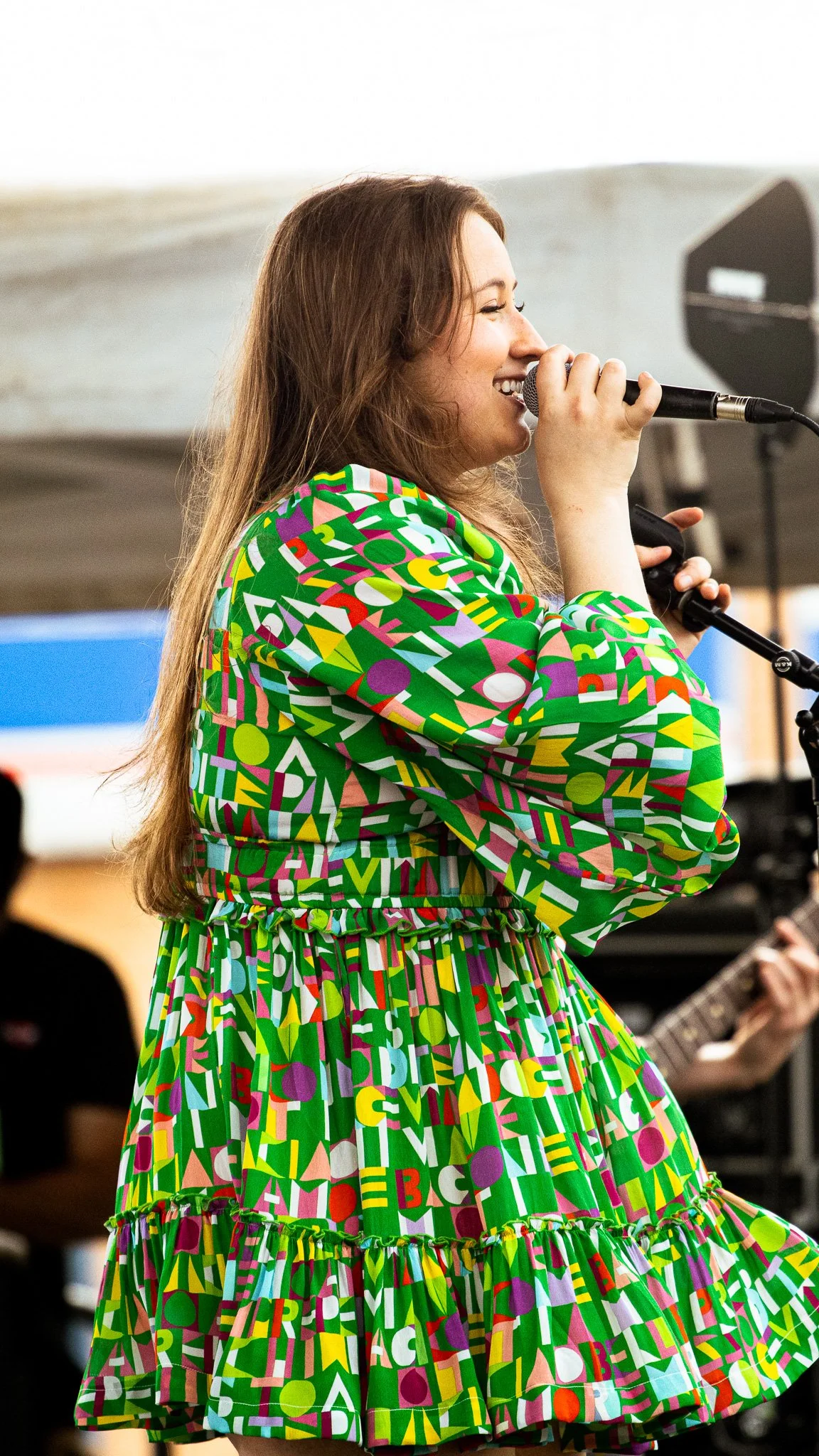 Woman singing into a microphone, wearing a colorful dress with letters and geometric shapes, with a person playing guitar in the background.