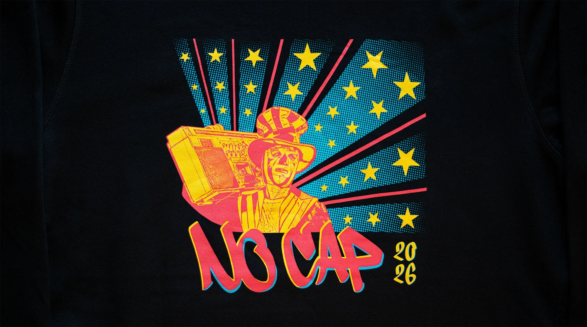 A colorful graphic design on black fabric showcasing a stylized figure of a woman wearing a hat and jacket, carrying a boombox on her shoulder, with yellow stars and blue dotted backgrounds radiating outward.