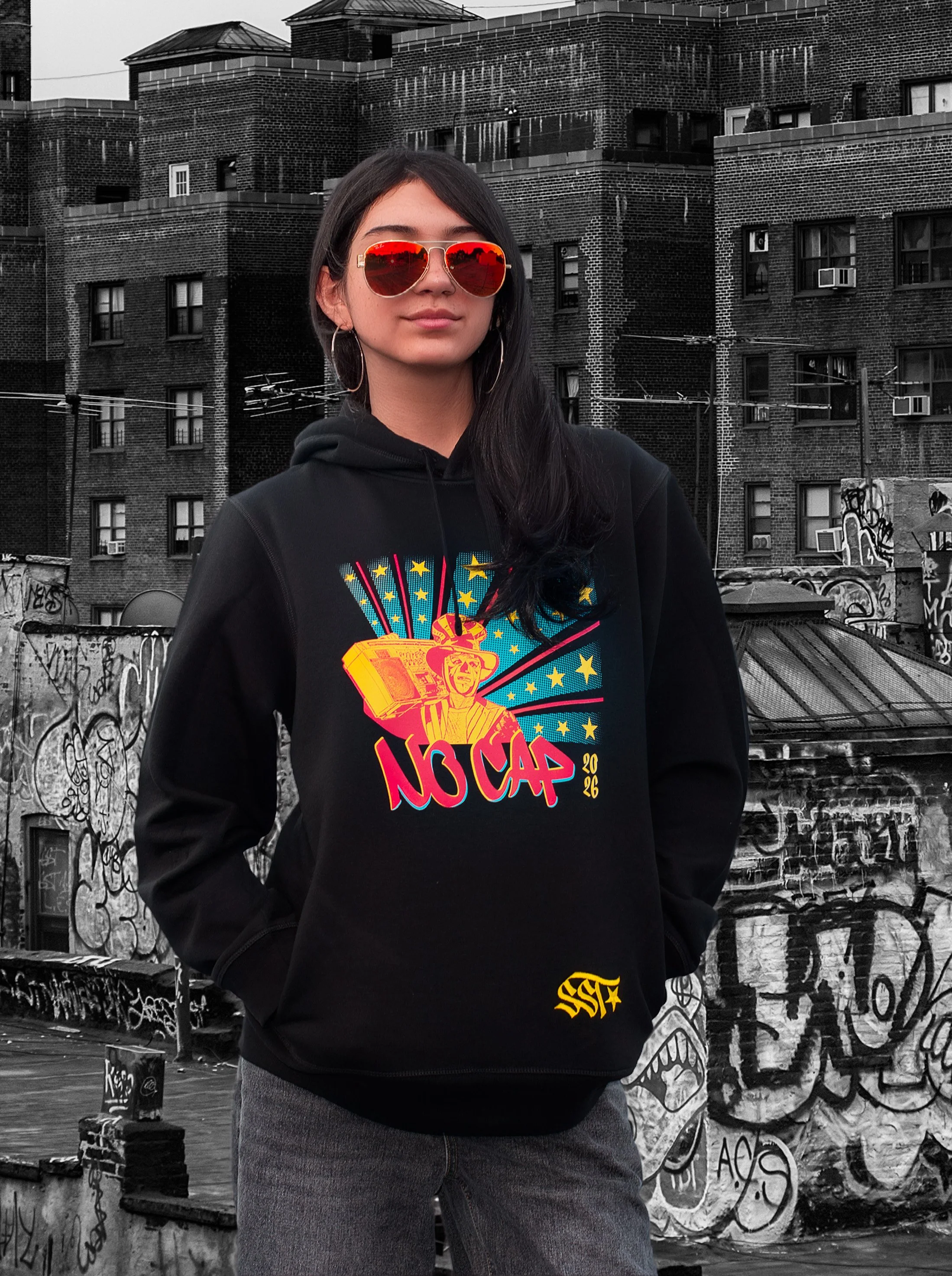 A woman wearing red sunglasses wears a limited edition black hoodie featuring the vibrant tricolor No Cap 2026 design. Behind her is a graffitied urban brick building background.