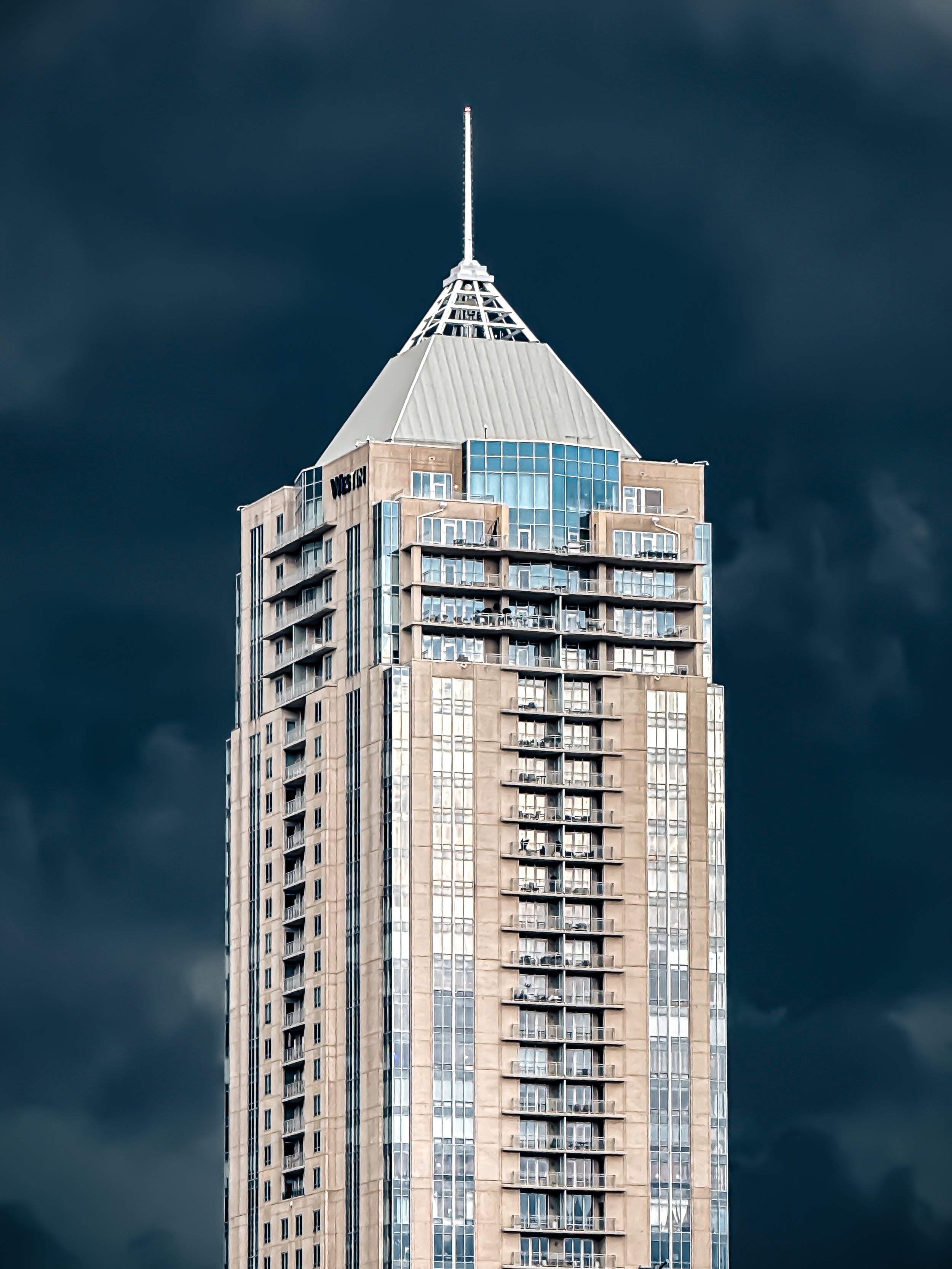 Tall skyscraper with glass windows and beige concrete exterior against a dark cloudy sky.