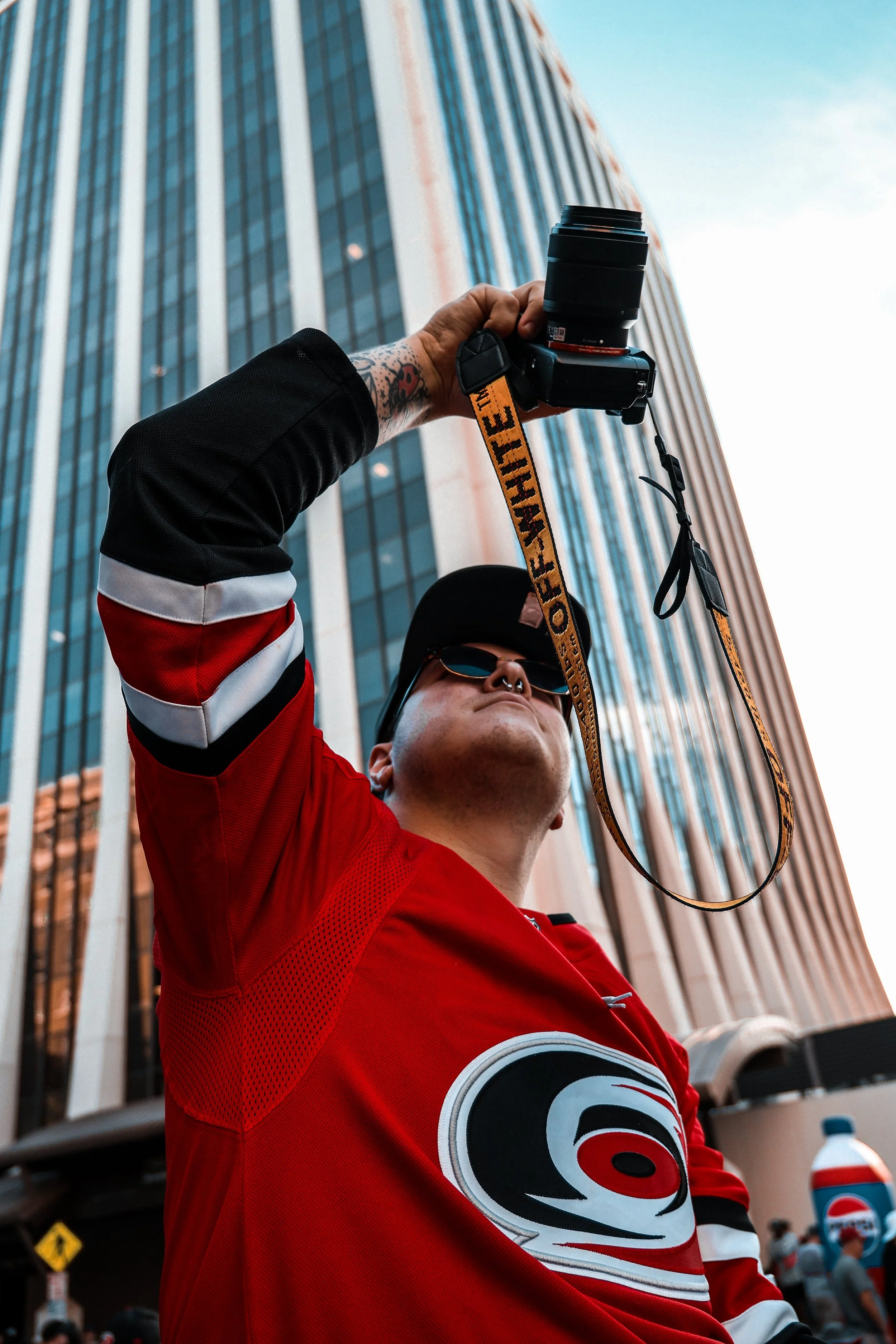 A person in a red sports jersey holding a camera in front of a tall glass building.