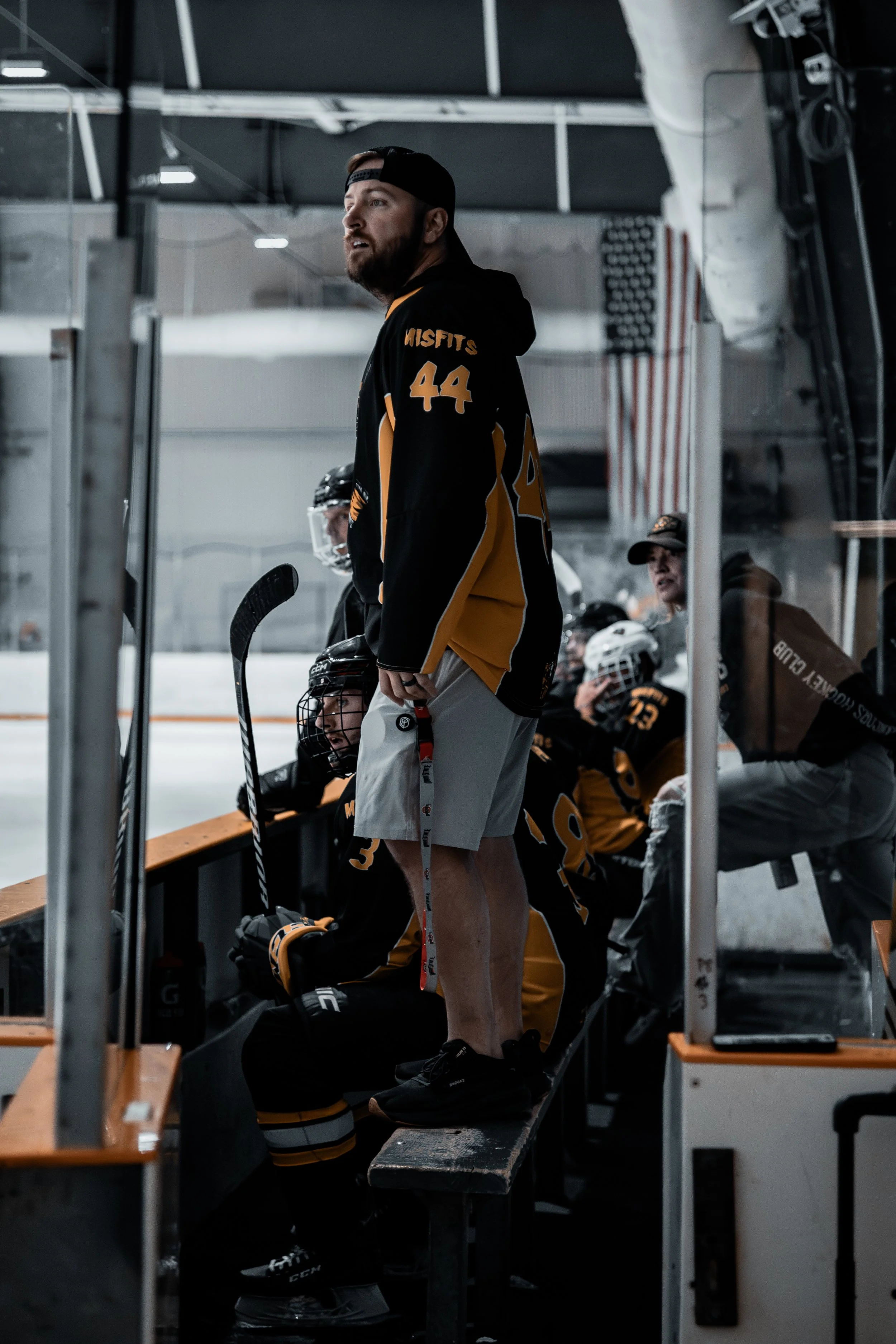 Hockey players sitting on the bench during a game, wearing black and yellow jerseys, some with helmets, in an indoor ice rink.